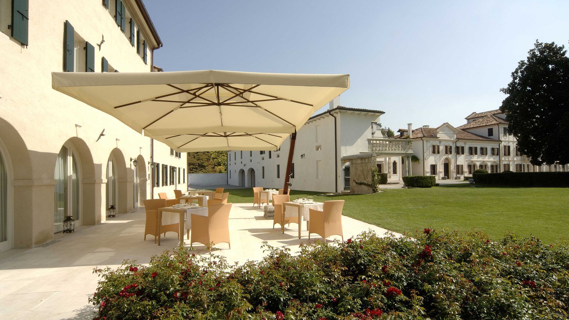 Zona pranzo all'aperto in un edificio bianco con tavoli, sedie e grandi ombrelloni.