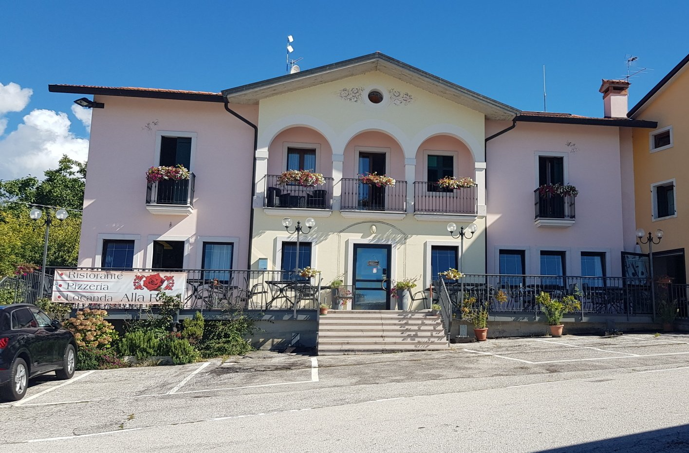 Edificio rosa e giallo con balcone ad arco, fiori, porta blu, giornata di sole.