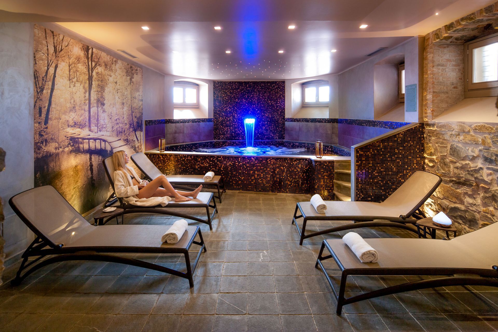 Una spa con una donna sdraiata su una sedia a sdraio. Luci blu illuminano una vasca idromassaggio e una cascata.