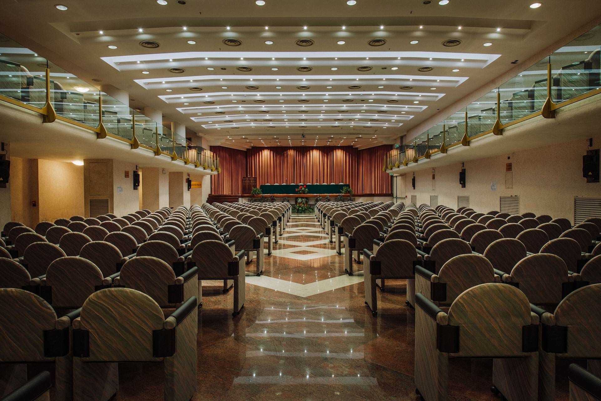 Auditorium con file di sedie di fronte a un palco con tende rosse. Illuminazione dall'alto.
