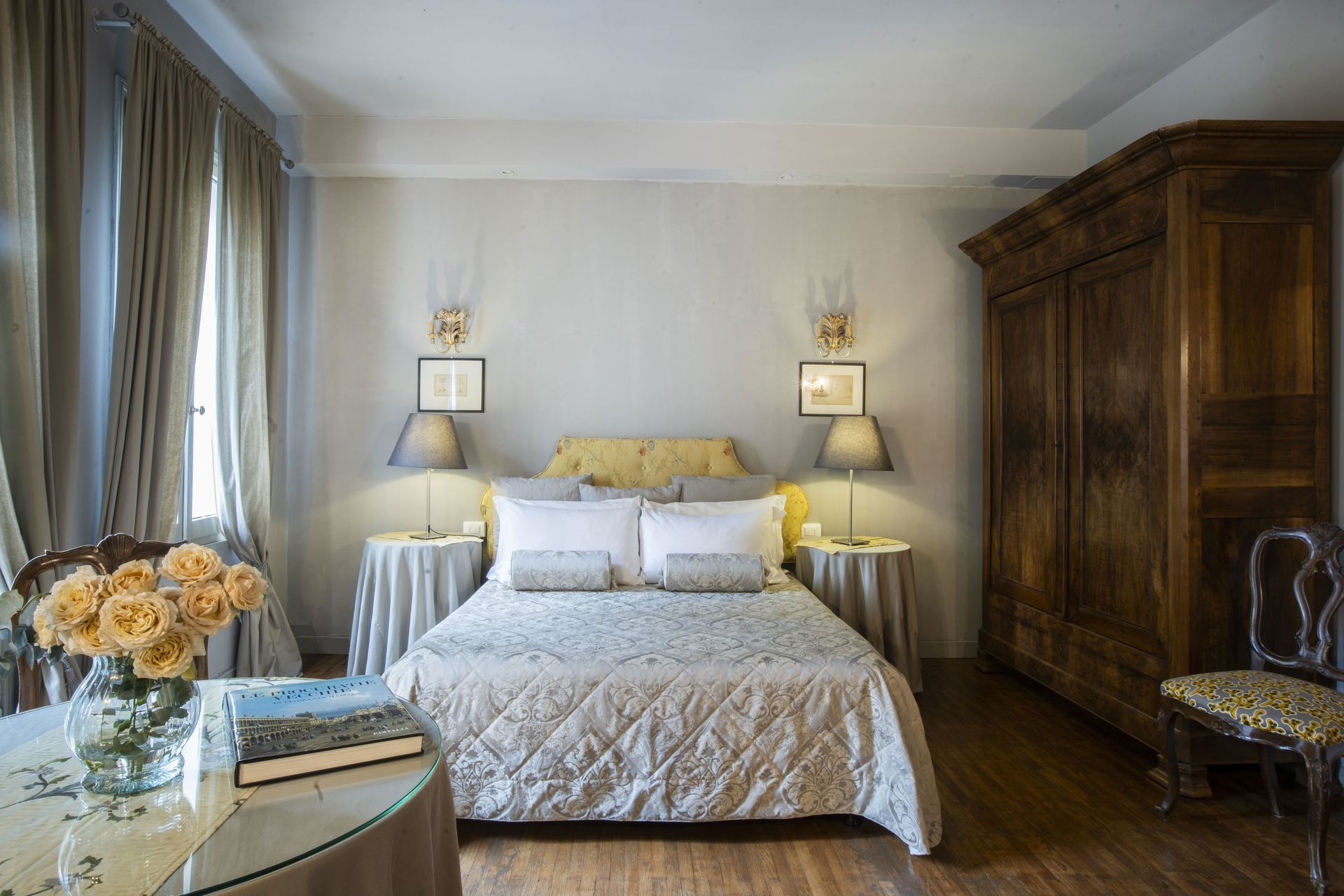 Elegante camera da letto: letto con trapunta, lampade, armadio antico, tavolo con fiori. Colori neutri, pavimento in legno.