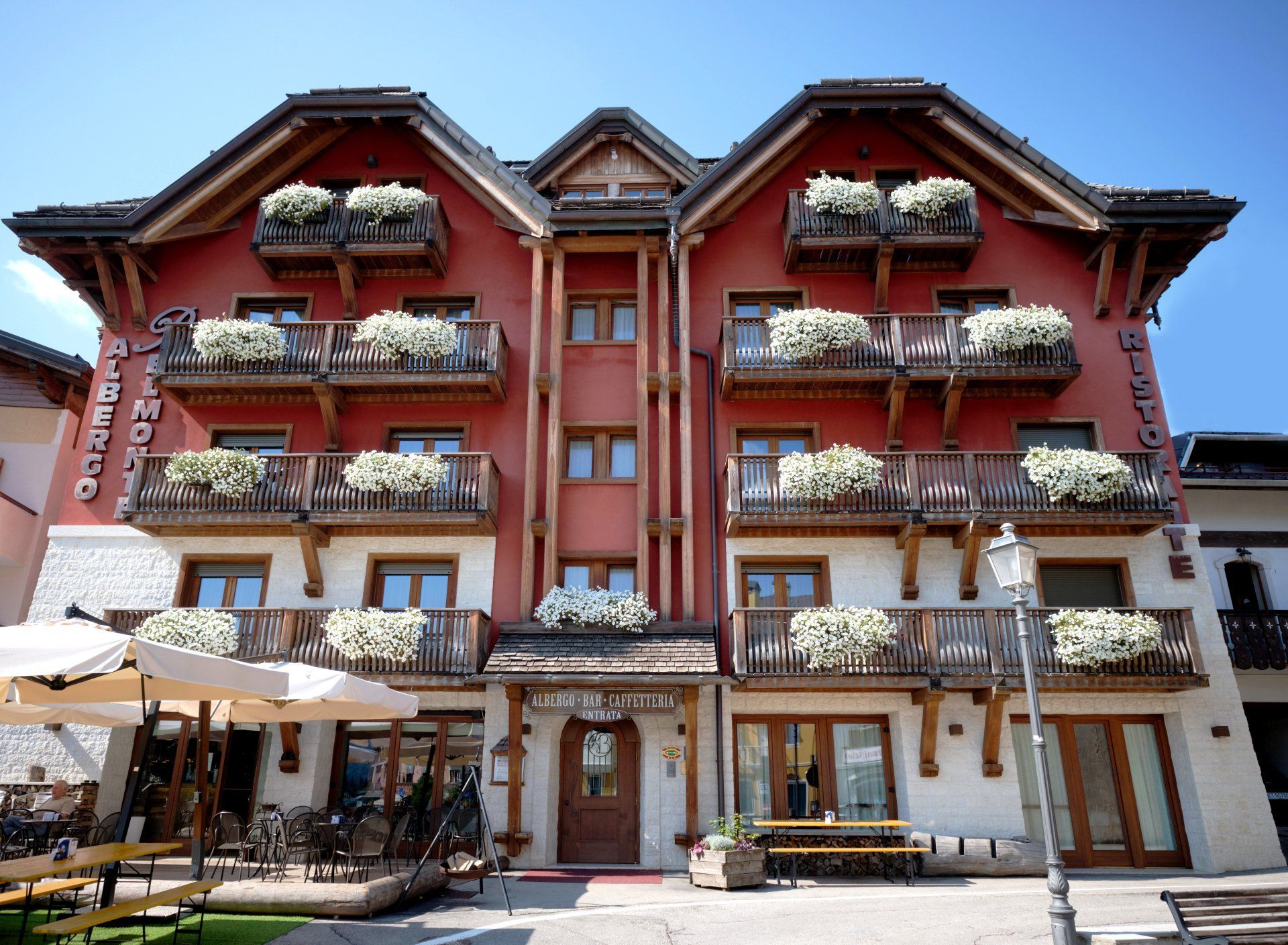 Esterno dell'hotel, facciata rossa, balconi in legno con fiori, posti a sedere all'aperto.