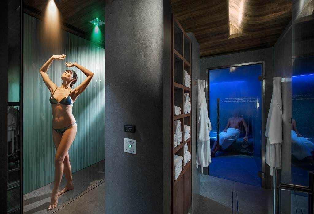 Donna sotto la doccia di una spa, con le braccia alzate, accanto a una colonna grigio scuro.