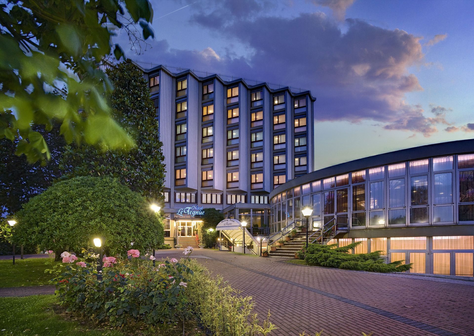 Esterno dell'hotel al tramonto, cielo azzurro, finestre illuminate, sentiero in pietra, vegetazione.