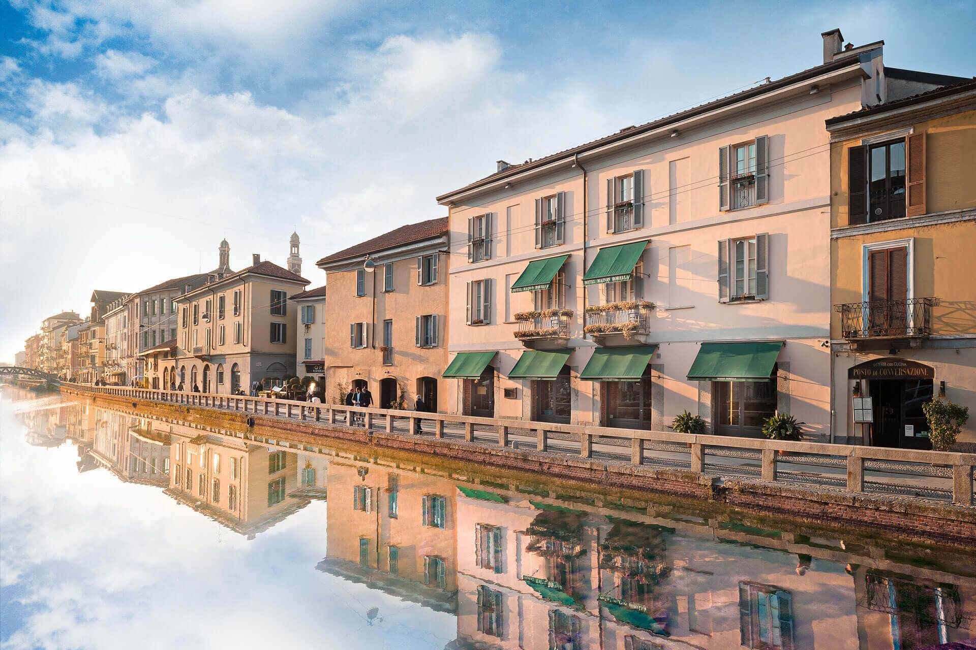 Edifici lungo un canale a Milano, riflessi nell'acqua. Strutture beige con tende da sole verdi, sotto un cielo azzurro.