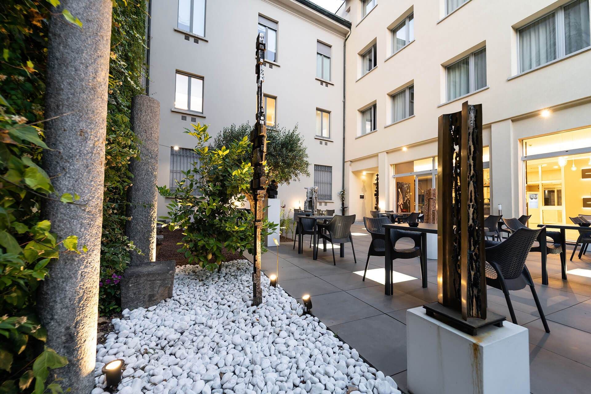 Ristorante nel cortile con tavoli, sedie e opere d'arte, circondato da un sentiero in pietra bianca e da edifici.