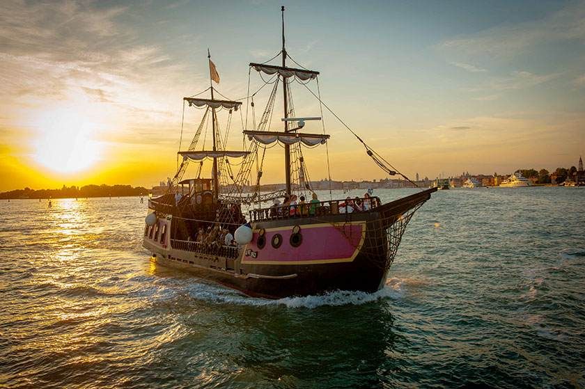 Nave pirata che naviga sull'acqua al tramonto, con dettagli rosa sullo scafo, persone sul ponte.