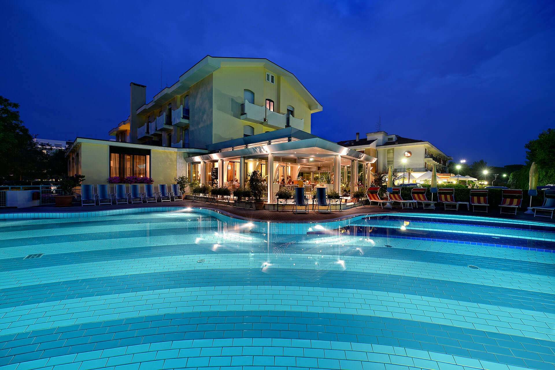 Vista notturna di un hotel con piscina illuminata. Edificio giallo, acqua blu, posti a sedere all'aperto visibili.