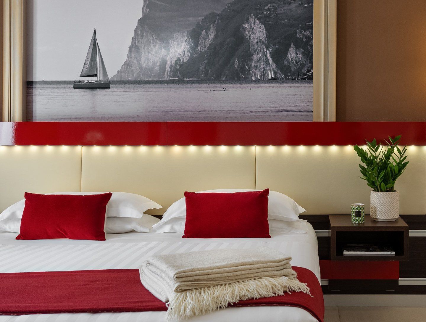 Camera da letto con dettagli rossi, tra cui cuscini e finiture della testiera, e una stampa artistica di una barca a vela.