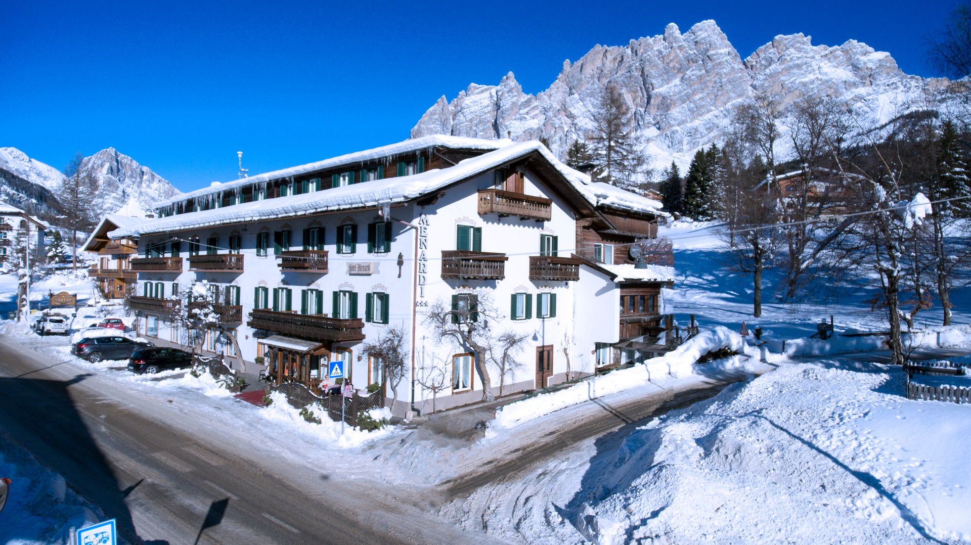 Hotel innevato sulle Dolomiti, montagne sullo sfondo. Edificio bianco con balconi marroni.