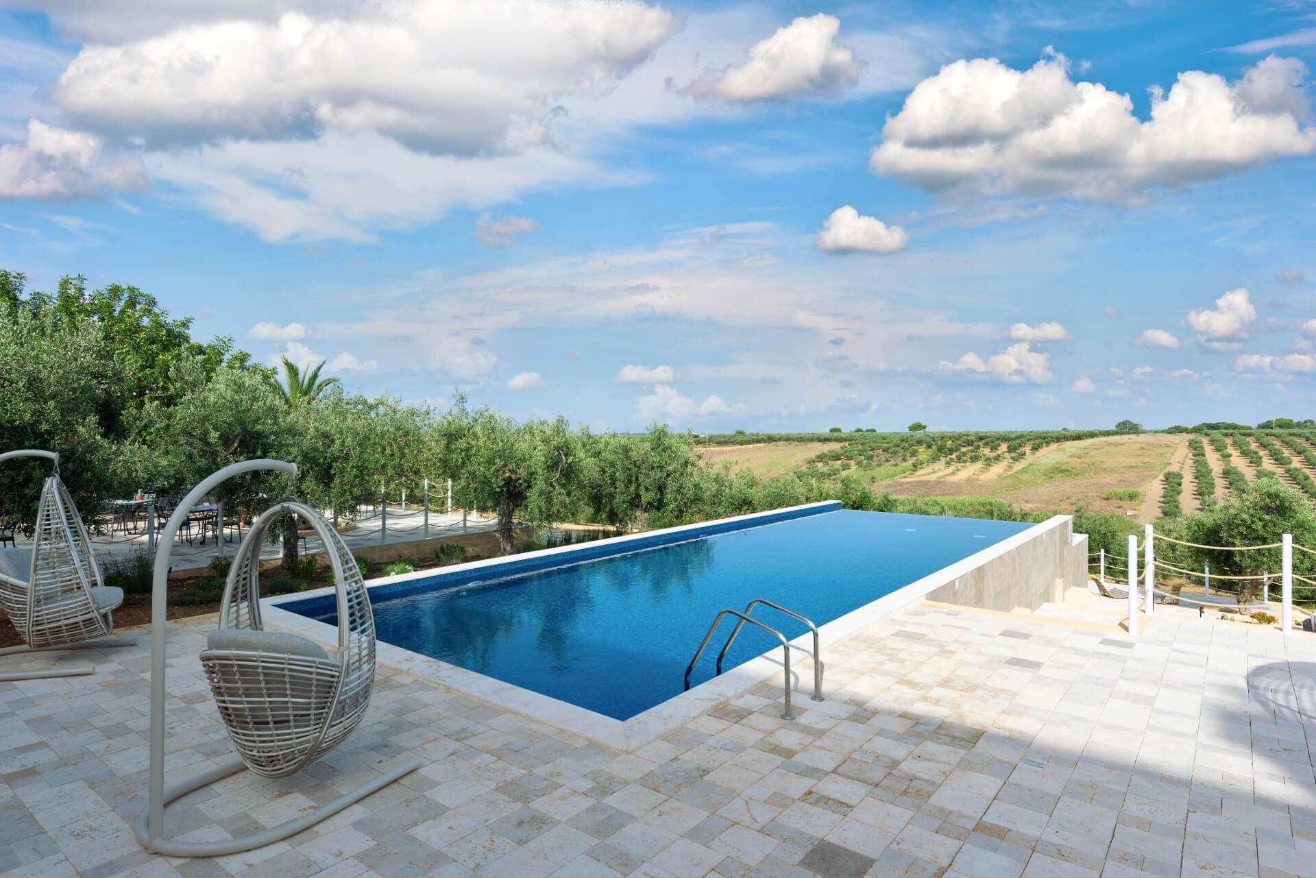 Piscina a sfioro con vista su un paesaggio lussureggiante sotto un cielo azzurro, dotata di sedie sospese.