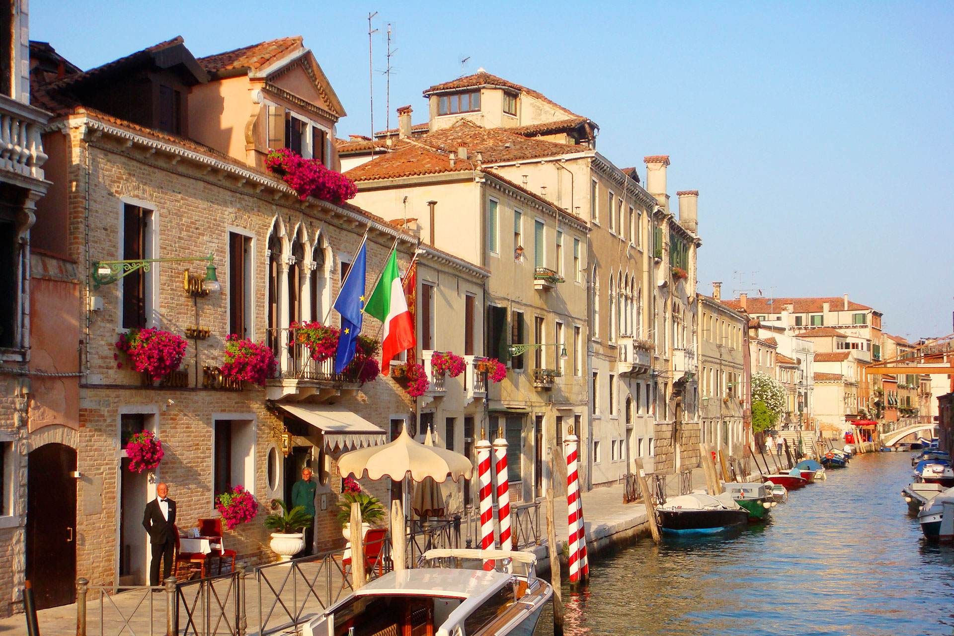 Canale di Venezia con edifici, barche e bandiera italiana; giornata di sole.