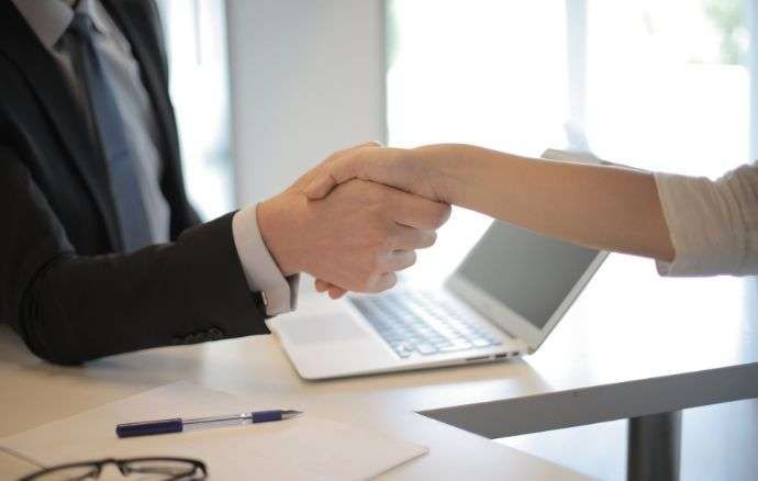 Due persone sedute a una scrivania si stringono la mano sopra un computer portatile, a simboleggiare un accordo.