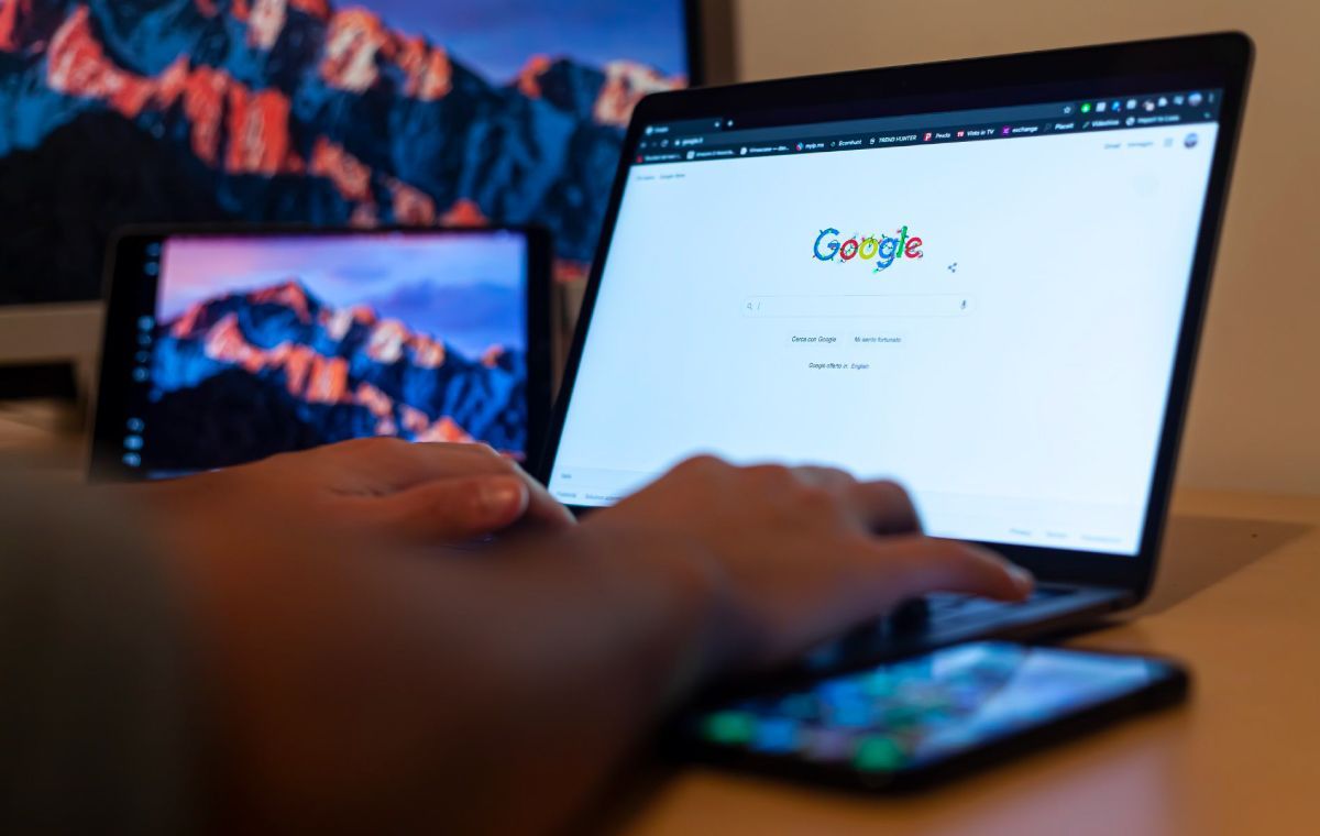 A person typing on a laptop with a Google search page open, with a tablet and smartphone nearby on a desk.