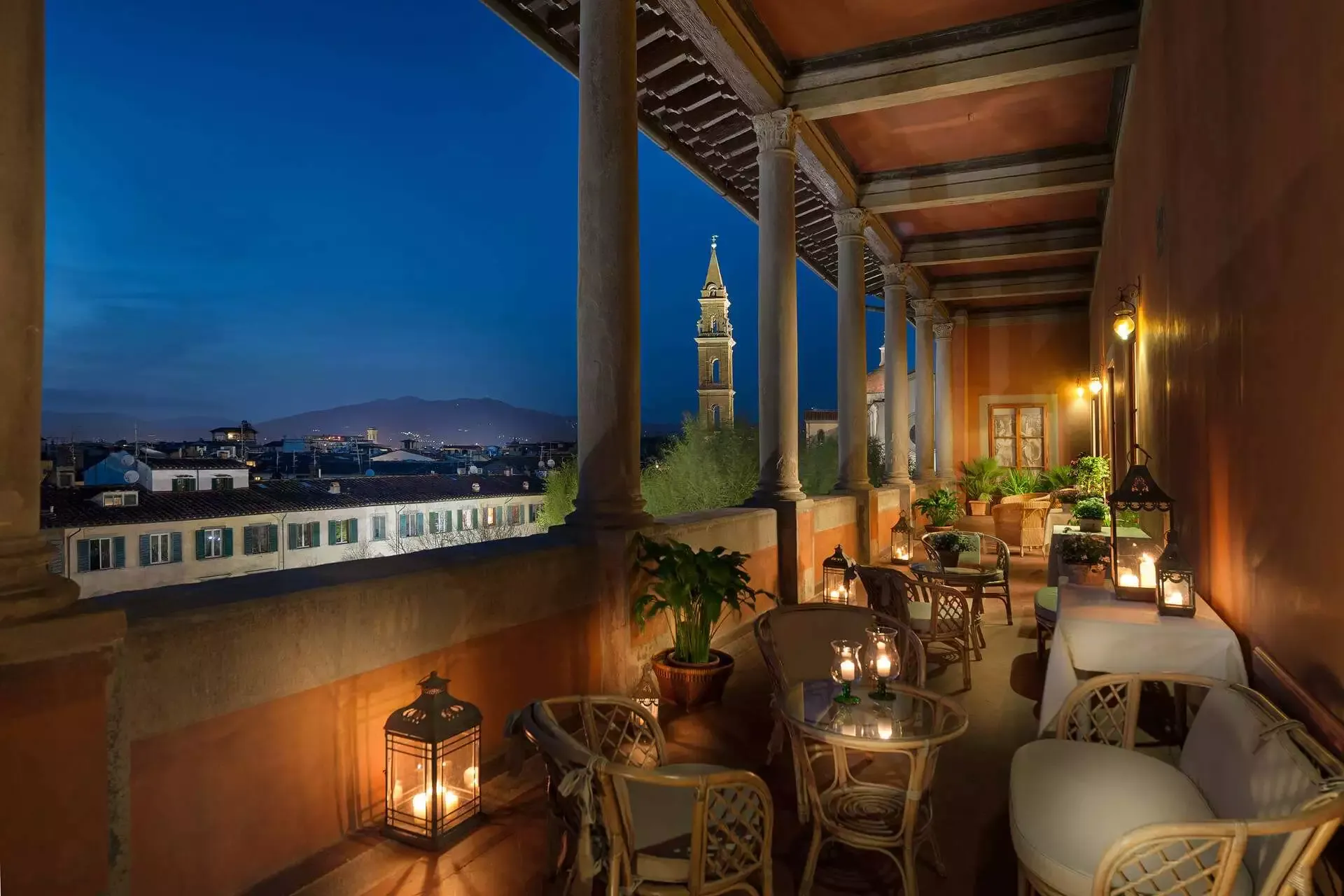 Bar con balcone e vista sulla città di notte; lanterne e tavoli illuminati, con un'alta torre in lontananza.