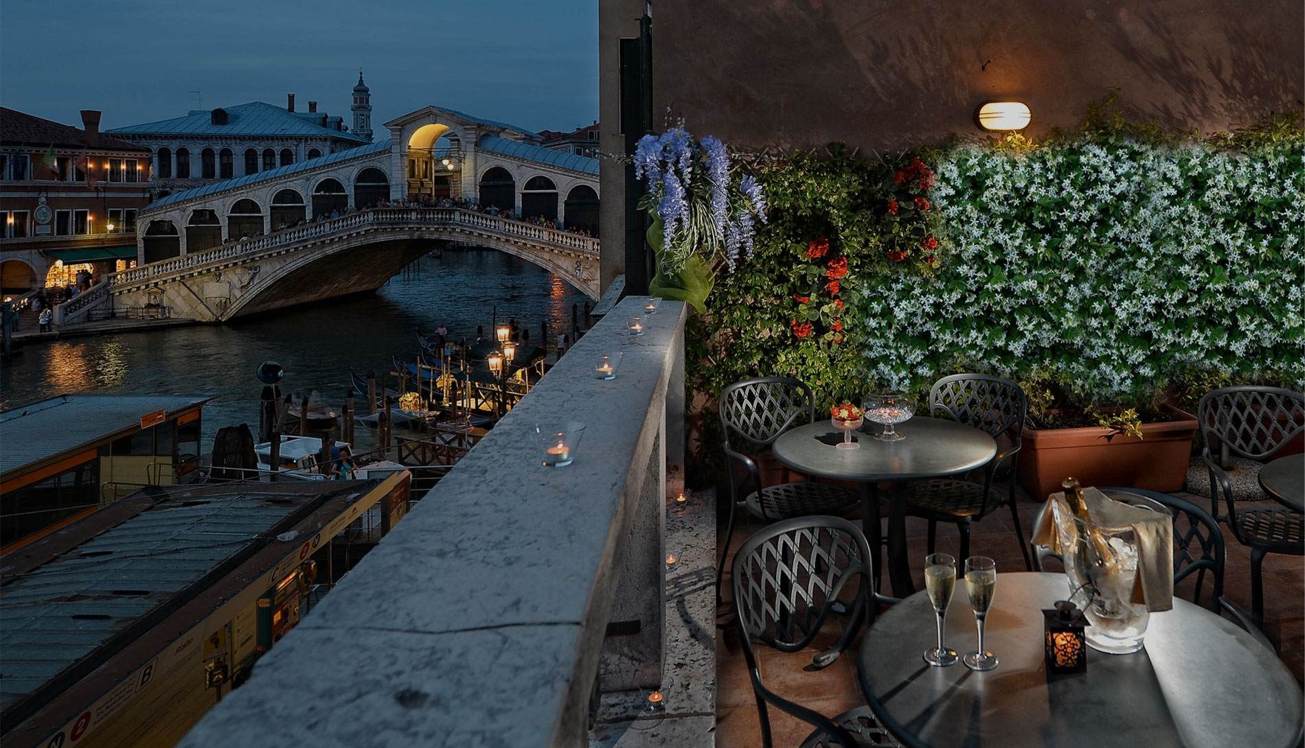 Patio del ristorante con vista sul Ponte di Rialto a Venezia al tramonto. Tavoli, sedie, fiori e il ponte gremito di gente.