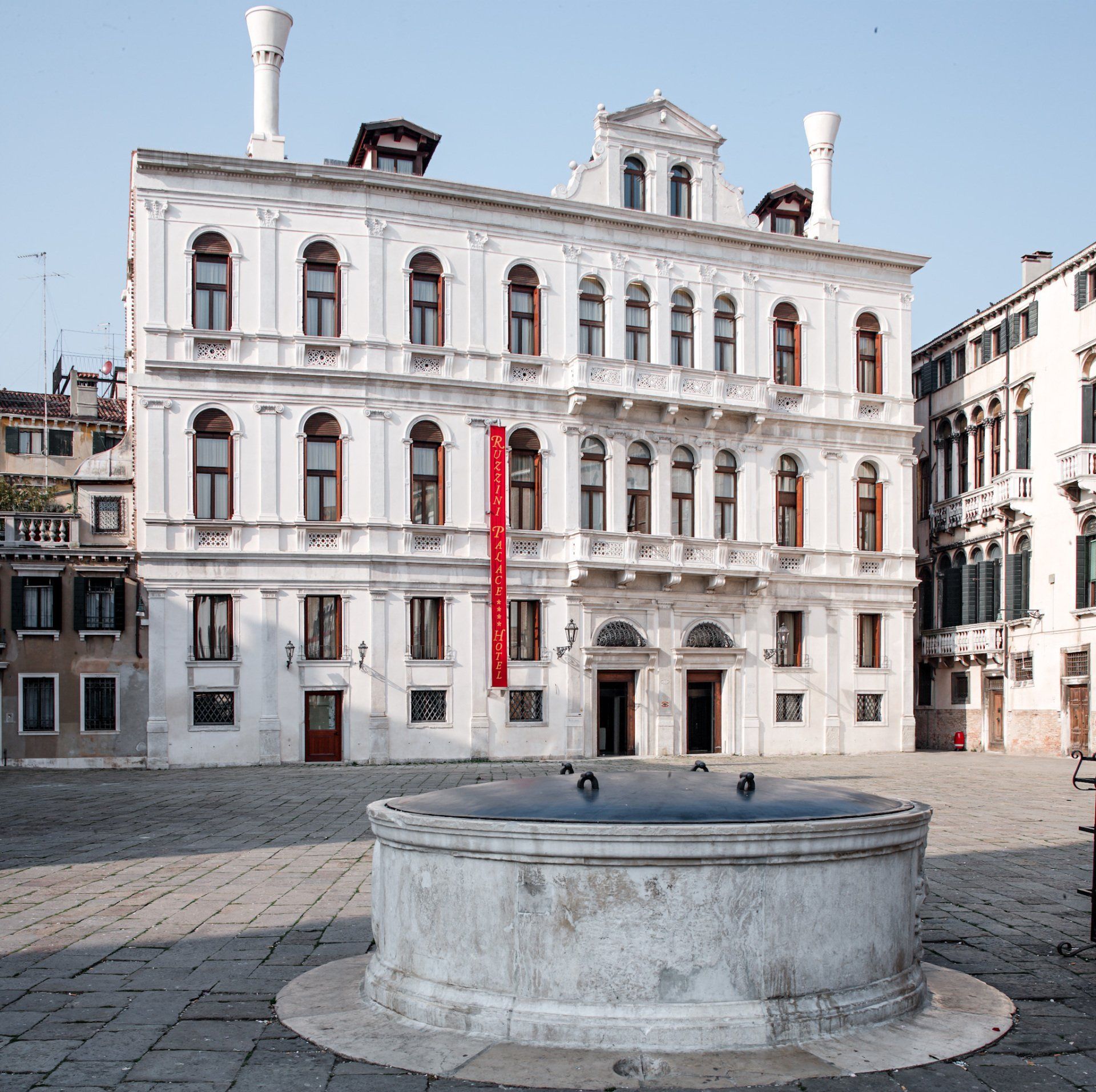 Edificio veneziano bianco con accenti rossi, una fontana e una piazza acciottolata.