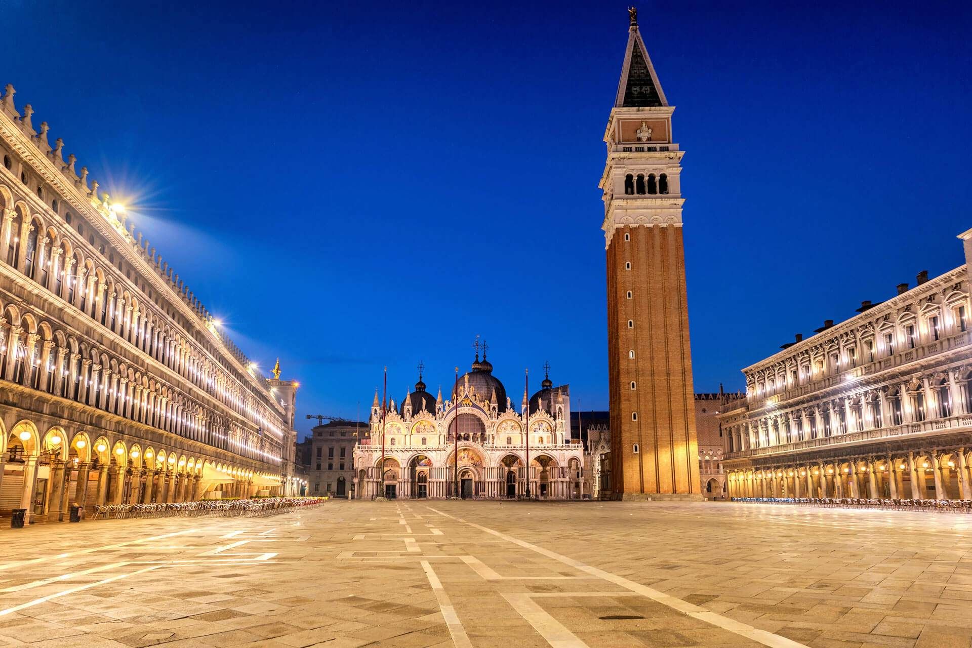 Piazza San Marco a Venezia al tramonto, con edifici illuminati, un alto campanile e un cielo blu scuro.