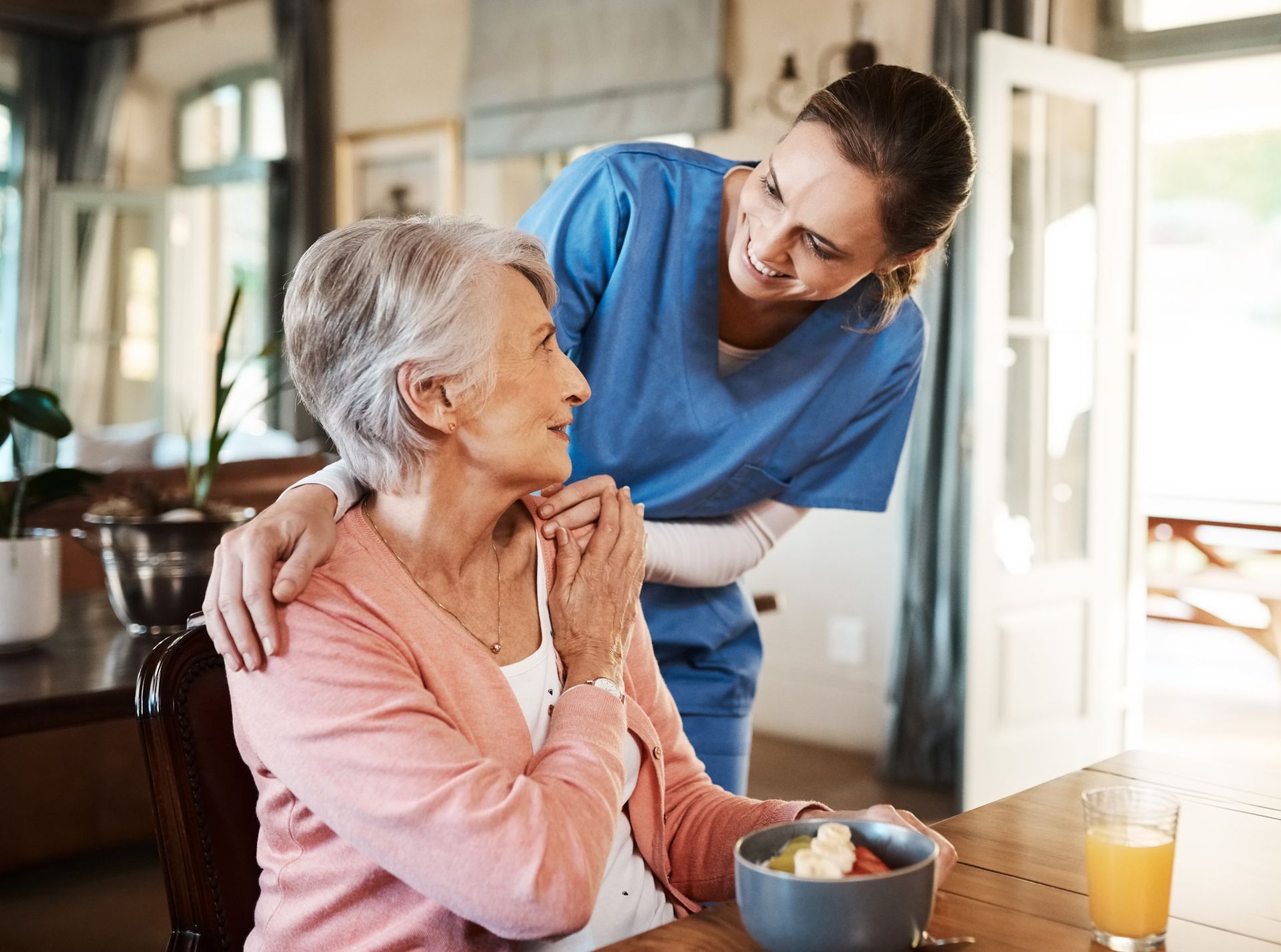 Caregiver in blue helping an elderly woman at a table with breakfast in a bright home kitchen