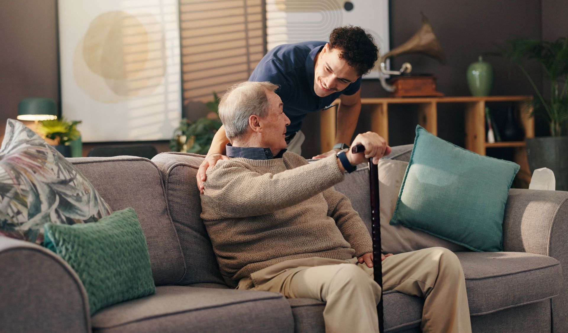 Caregiver helping an older adult stand from a sofa in a cozy living room