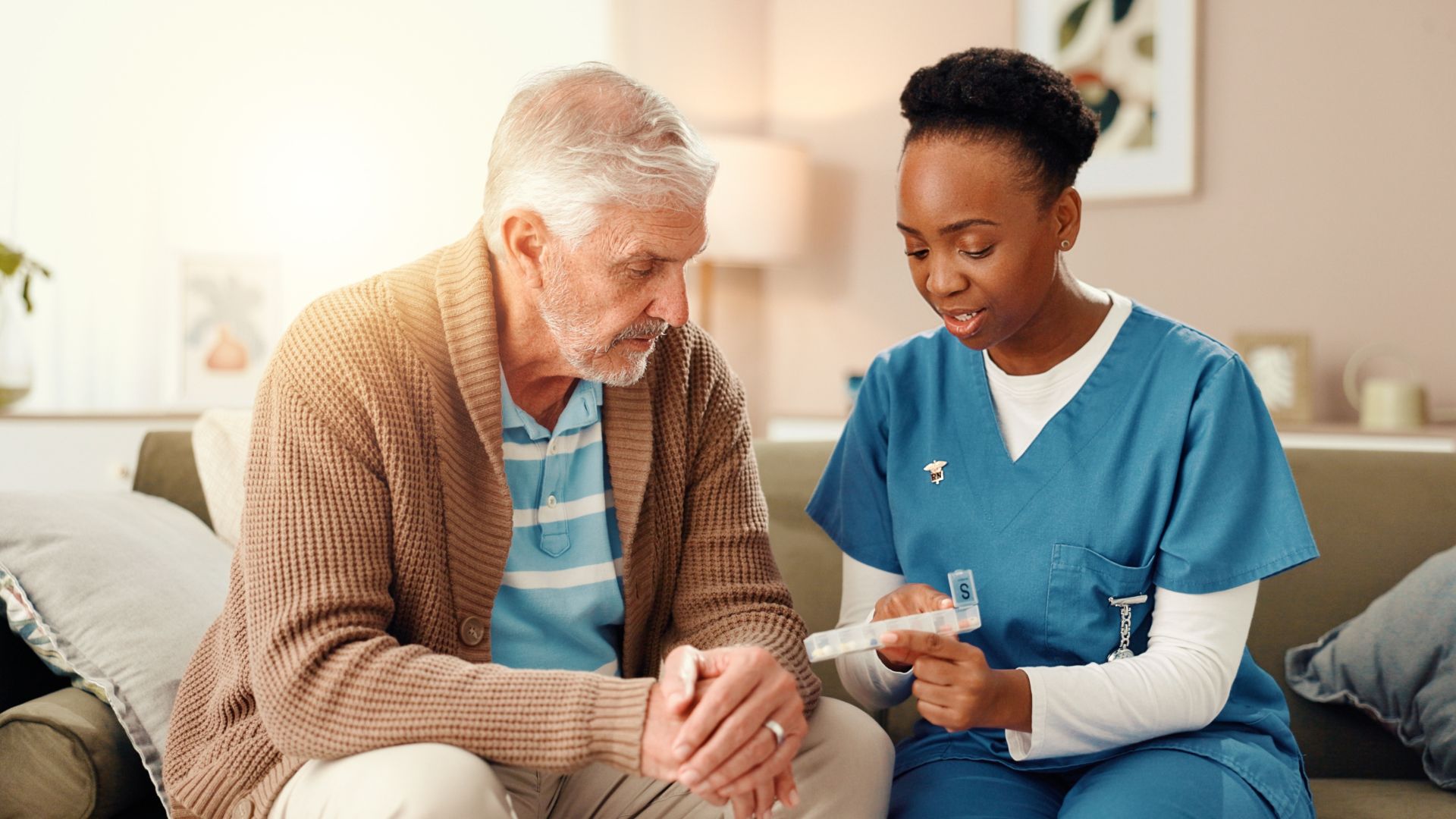Nurse in blue scrubs showing a tablet to a seated patient in a cozy home setting