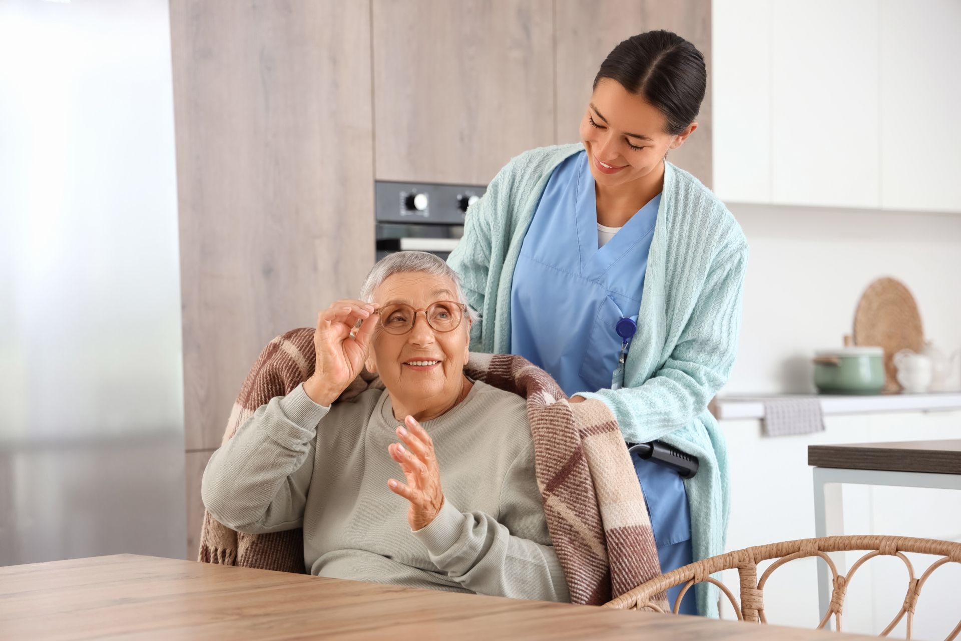 Caregiver helping an elderly man seated at a dining table in a bright kitchen