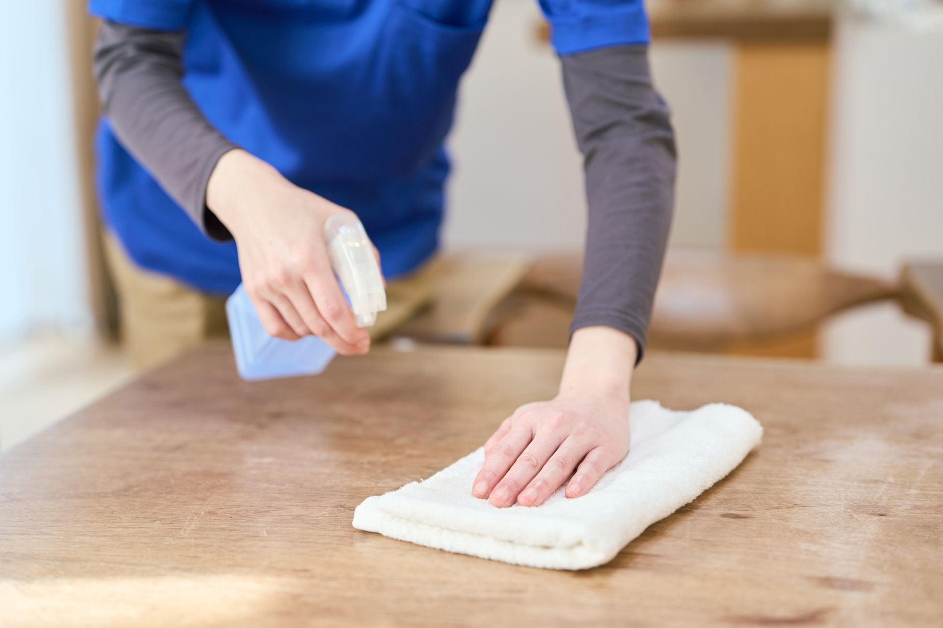 Person cleaning a wooden table with spray bottle and cloth in a home kitchen