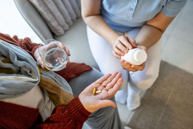 Person hands pills to another holding a water glass on a sofa
