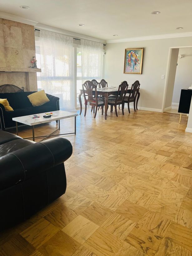 Bright living room with black sofa, glass coffee table, dining set, and parquet floor by large windows.