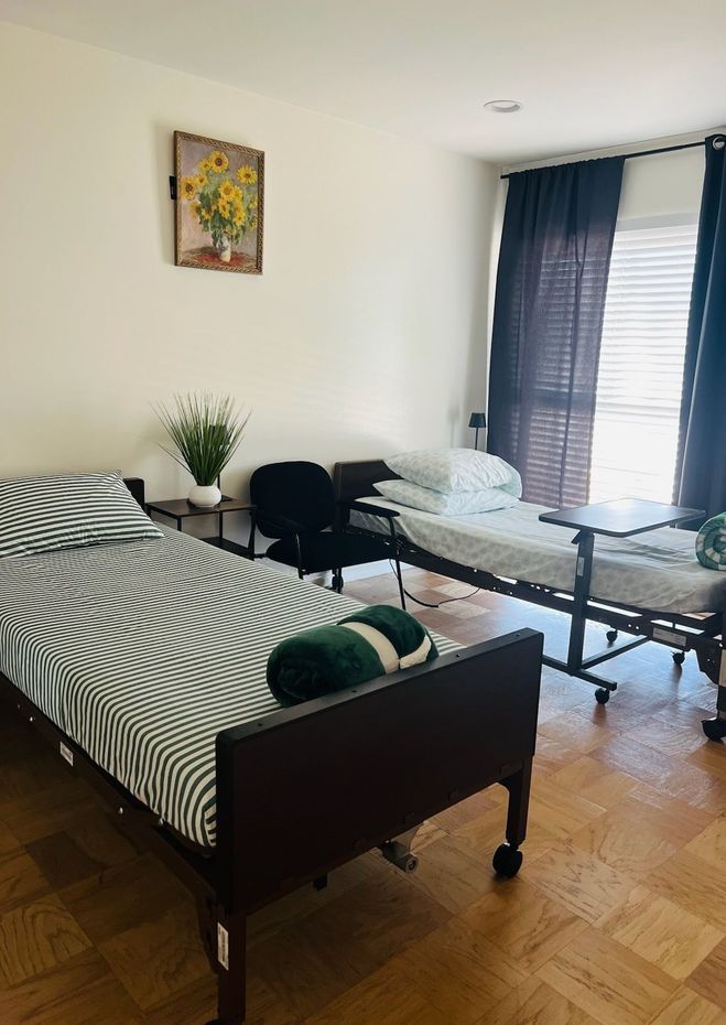 Two single beds in a bright room with a table, chairs, window blinds, and a painting on the wall