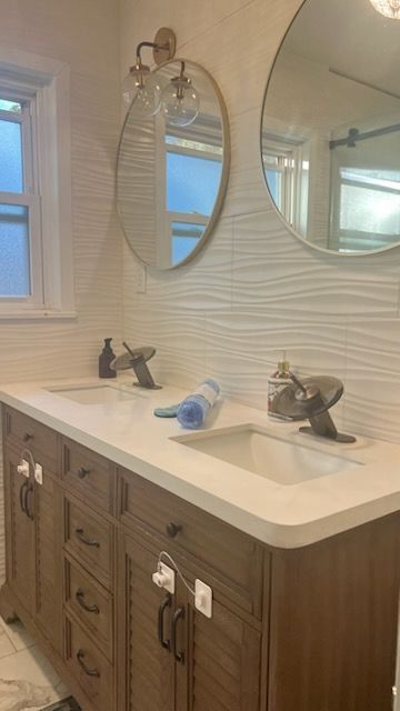 Modern bathroom vanity with white countertop, wood cabinets, twin sinks, and round mirrors beside a window