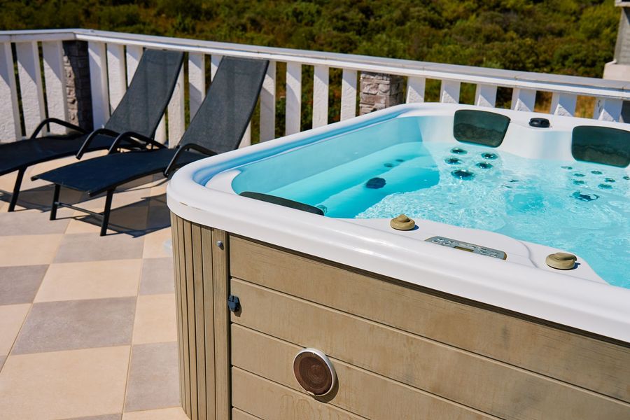 Hot tub on a sunny patio beside black lounge chairs and a wooden deck railing.