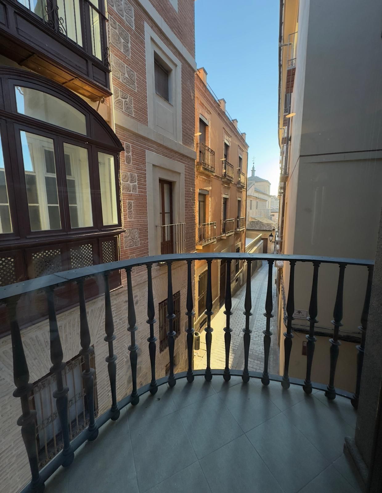 Vista desde un balcón curvo de hierro, en un ángulo elevado, de una calle estrecha e iluminada por el sol, entre edificios históricos de ladrillo.