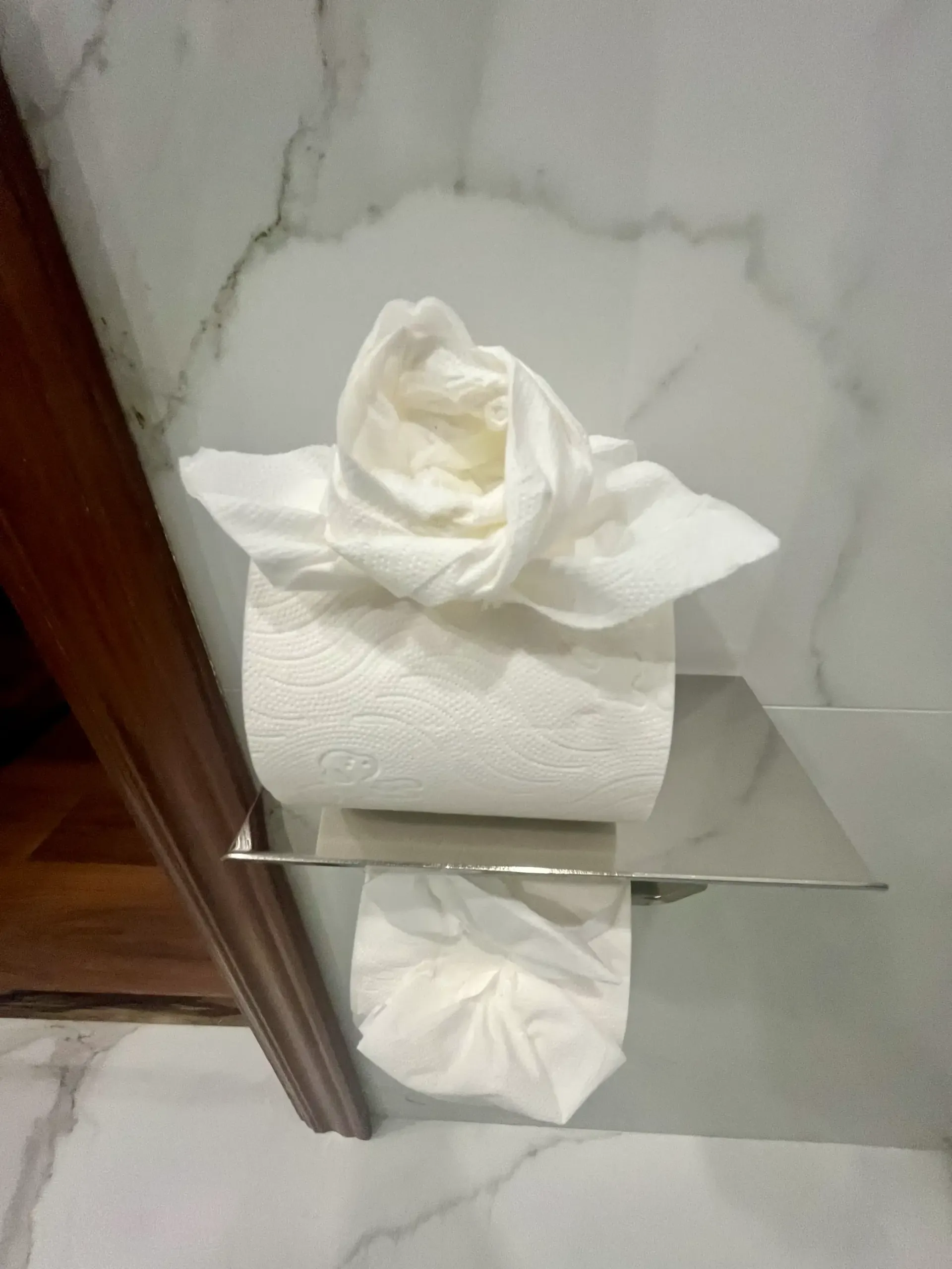 Un rollo de papel higiénico blanco con el extremo doblado en forma de flor decorativa, apoyado sobre un soporte cromado en un baño.