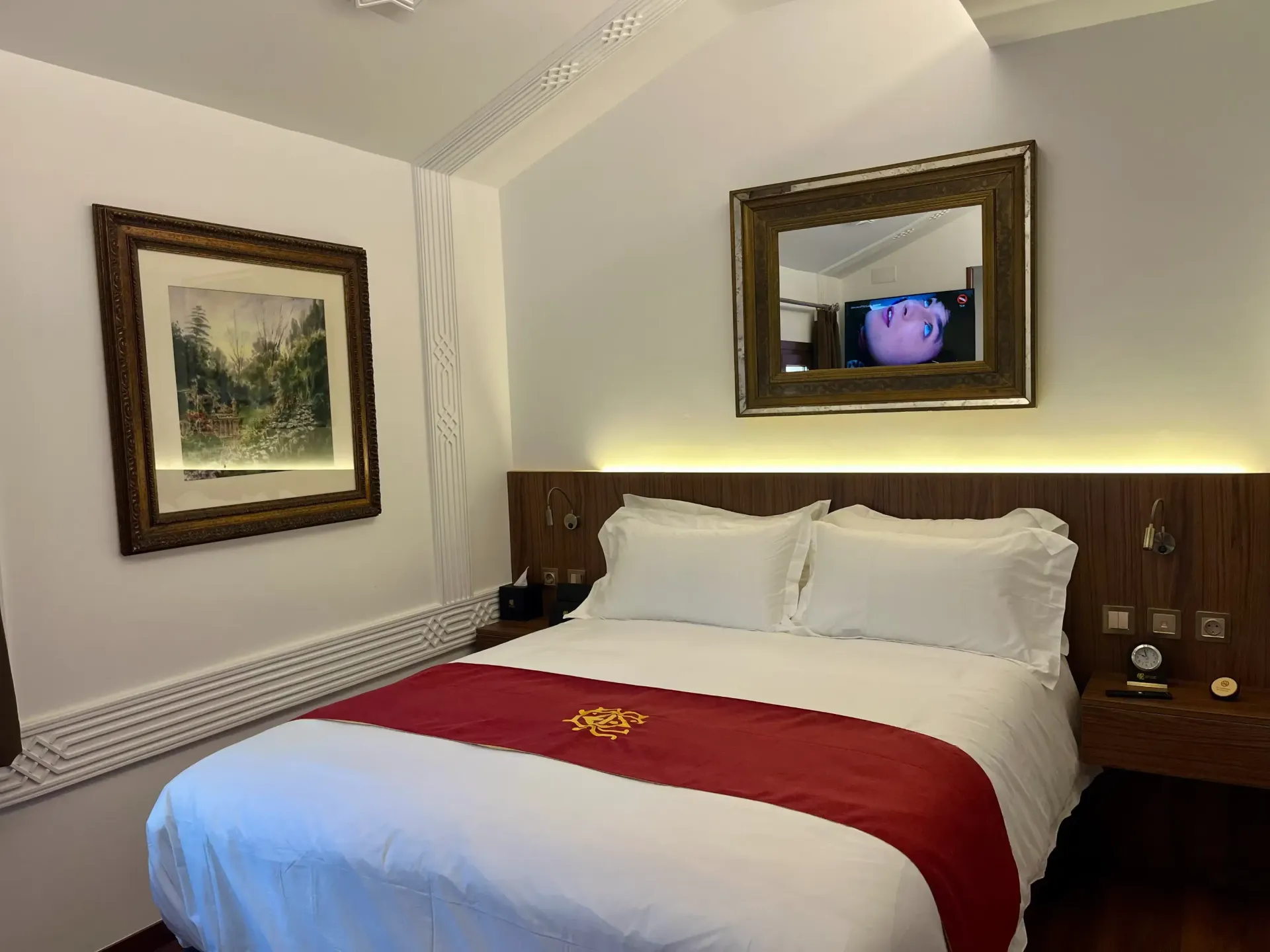 Una habitación de hotel con una cama tamaño queen, una alfombra roja, almohadas blancas, un cuadro con un paisaje enmarcado y un espejo sobre la cabecera.