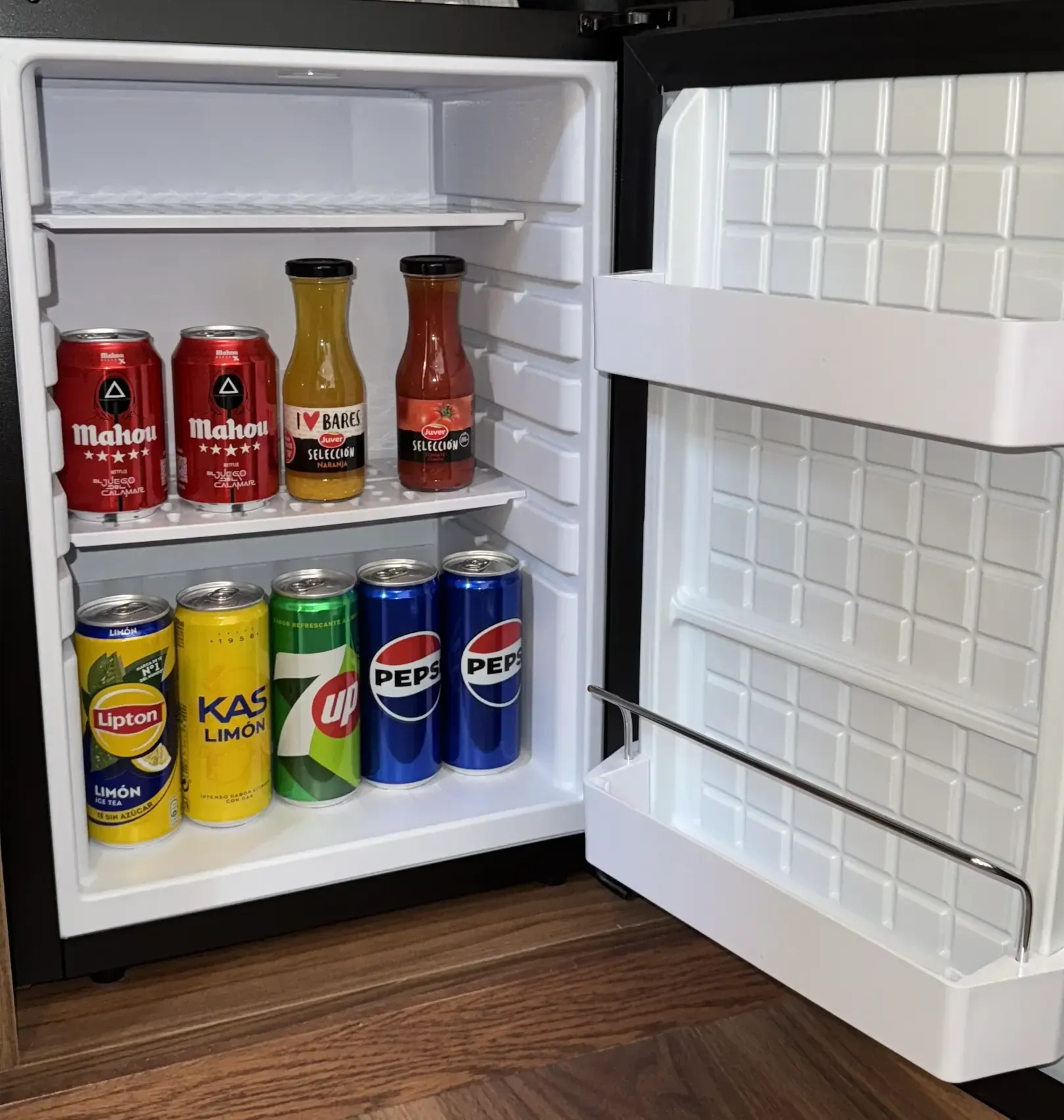 Un pequeño refrigerador que contiene varias latas y botellas de refrescos, jugos y cervezas, colocado sobre un piso de madera.