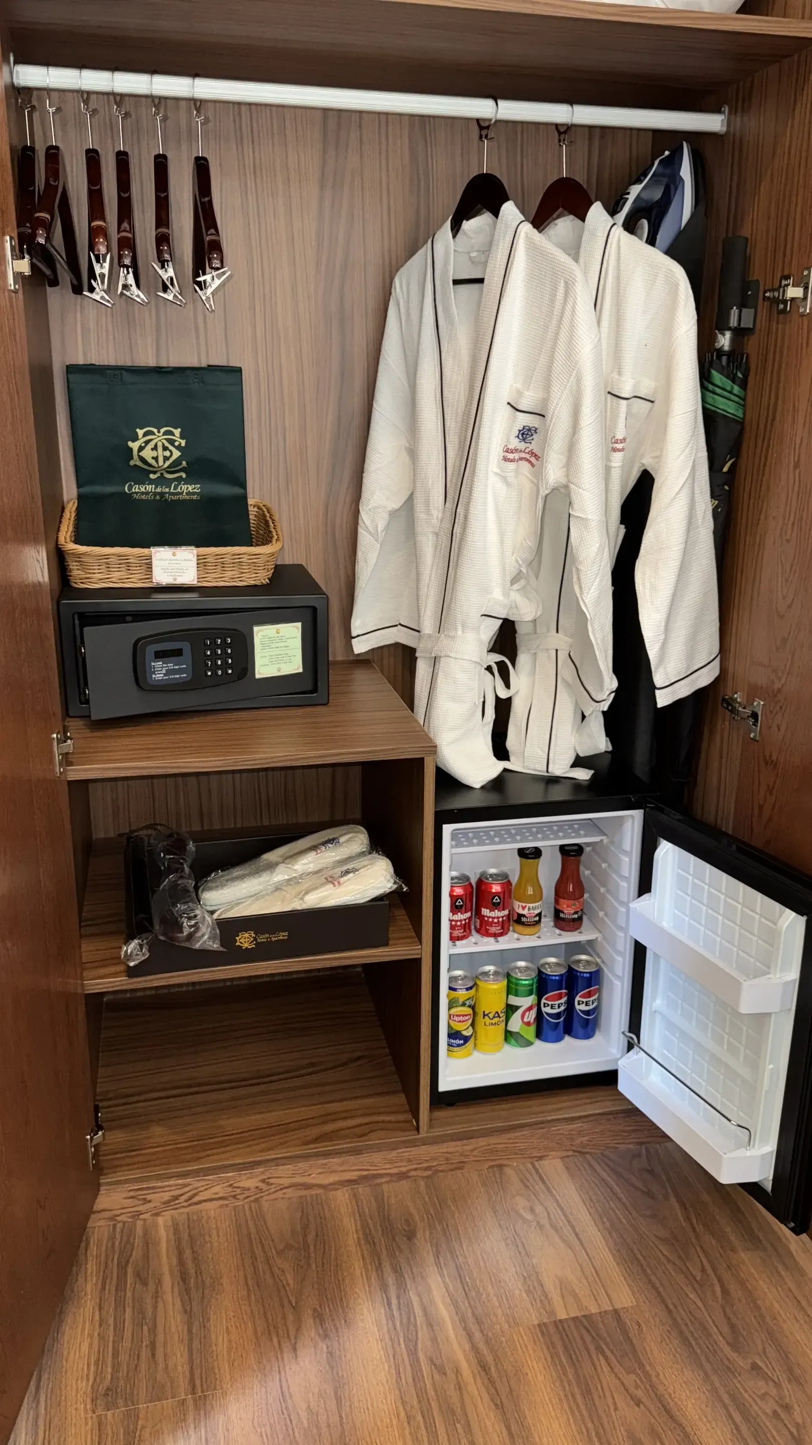 Un armario de hotel abierto que incluye una caja fuerte, dos albornoces blancos, estantes con zapatillas y un pequeño frigorífico.