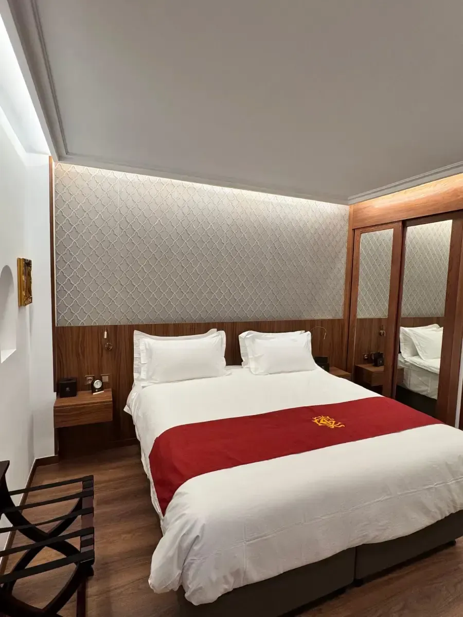 Una habitación de hotel moderna con una cama bien hecha, ropa de cama blanca, una alfombra granate y paredes revestidas de madera con espejos.
