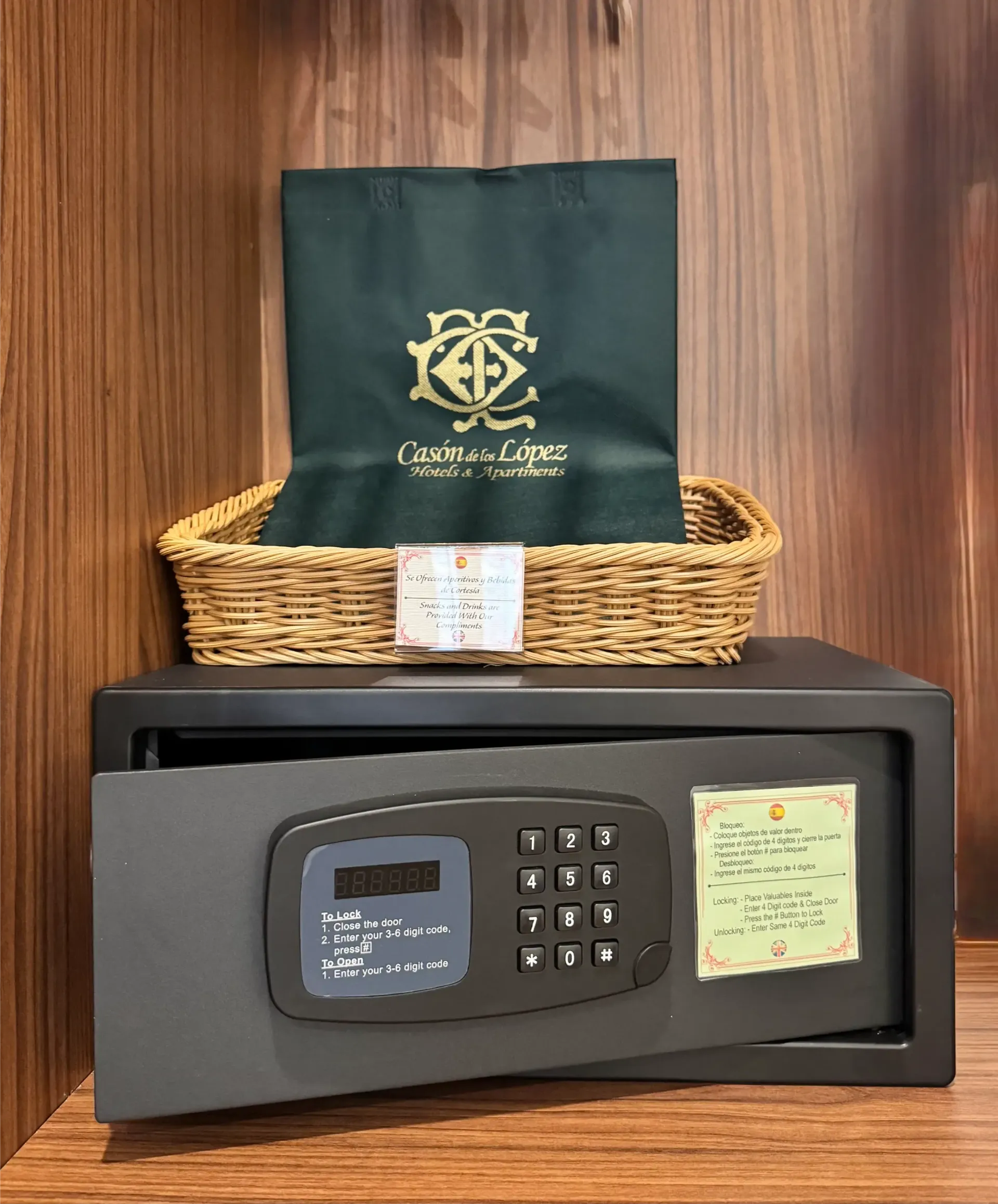 Una caja fuerte digital de hotel con la puerta abierta repisa sobre una estantería de madera, debajo de una cesta de mimbre que contiene una bolsa verde con el logotipo del hotel.