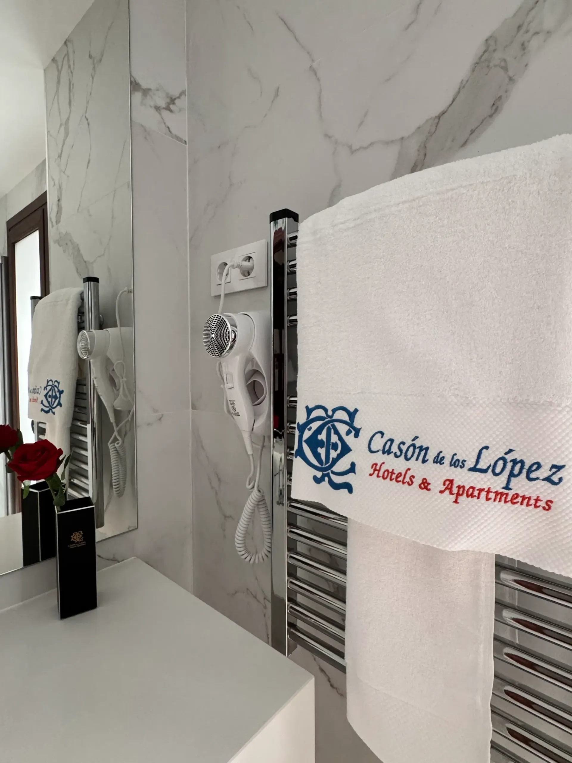 Un baño de hotel limpio, con paredes de mármol, un toallero con una toalla de marca, un secador de pelo y una sola rosa roja.