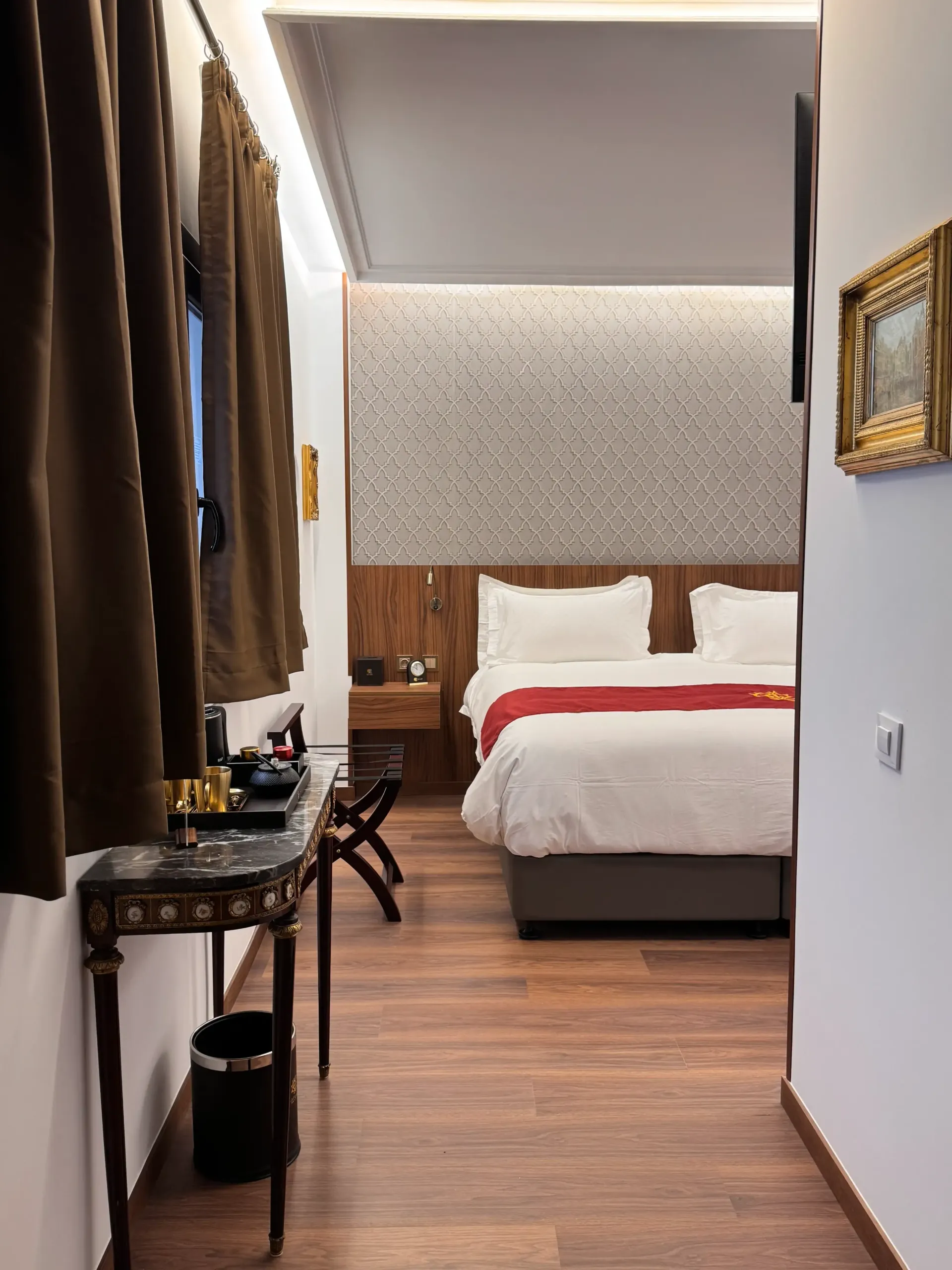 Una habitación de hotel con una cama con sábanas blancas y una manta roja, suelo de madera, cortinas oscuras y un cuadro enmarcado en la pared.