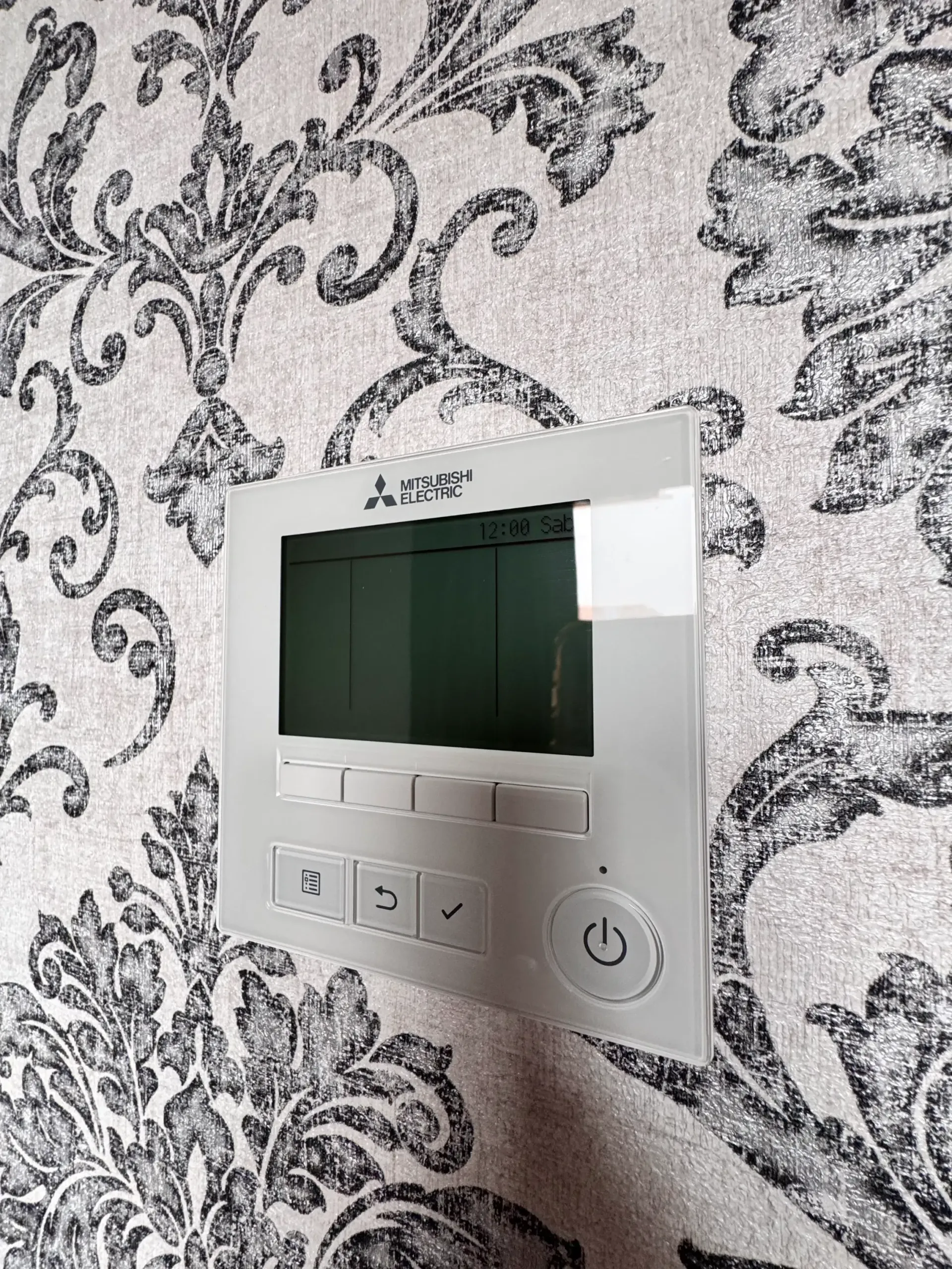 Un termostato de pared Mitsubishi con pantalla en blanco, montado en una pared con papel pintado de damasco blanco y negro.