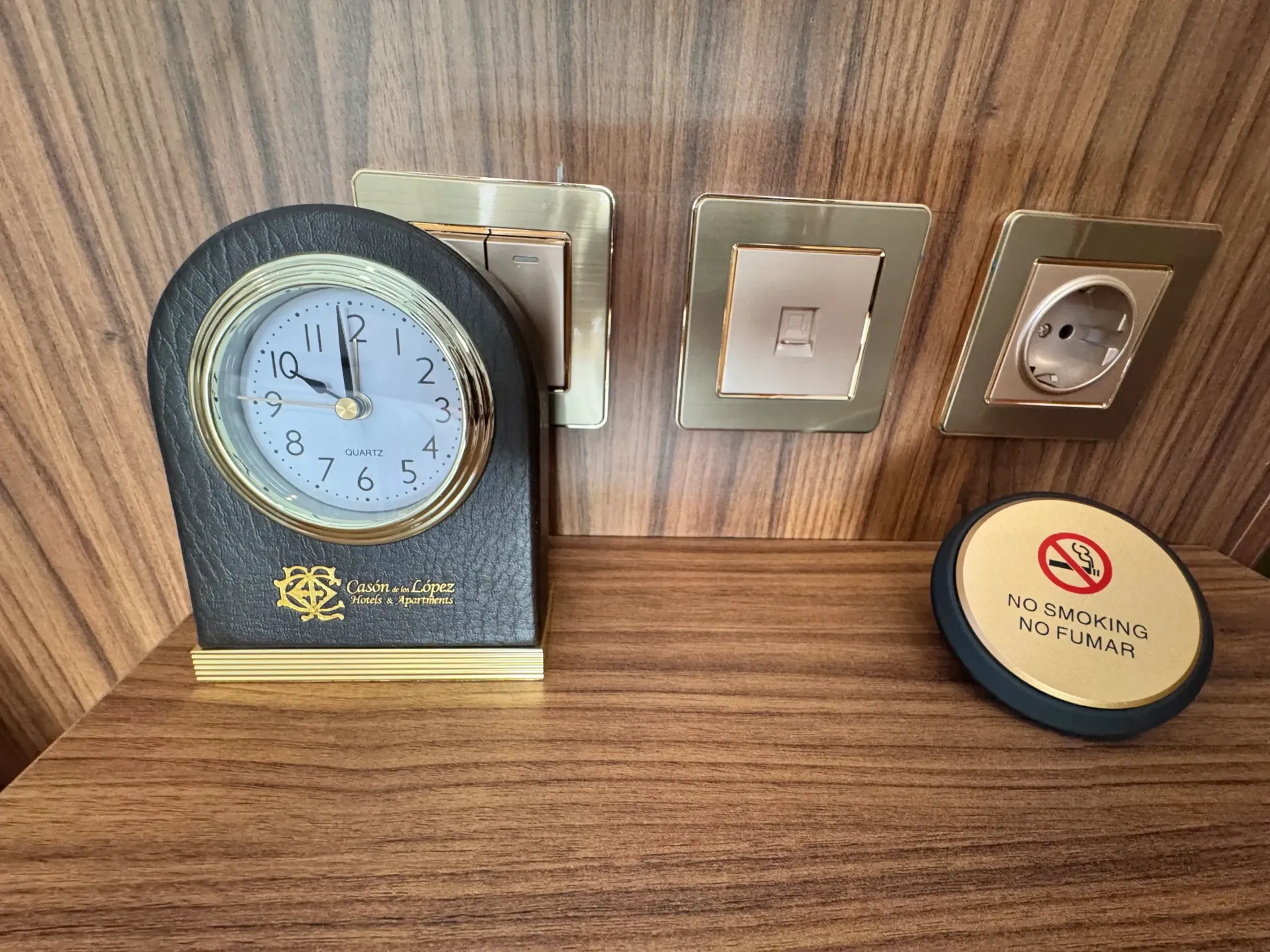 Un reloj analógico, interruptores de luz y una toma de corriente sobre una mesita de noche de madera, con un cartel redondo de 