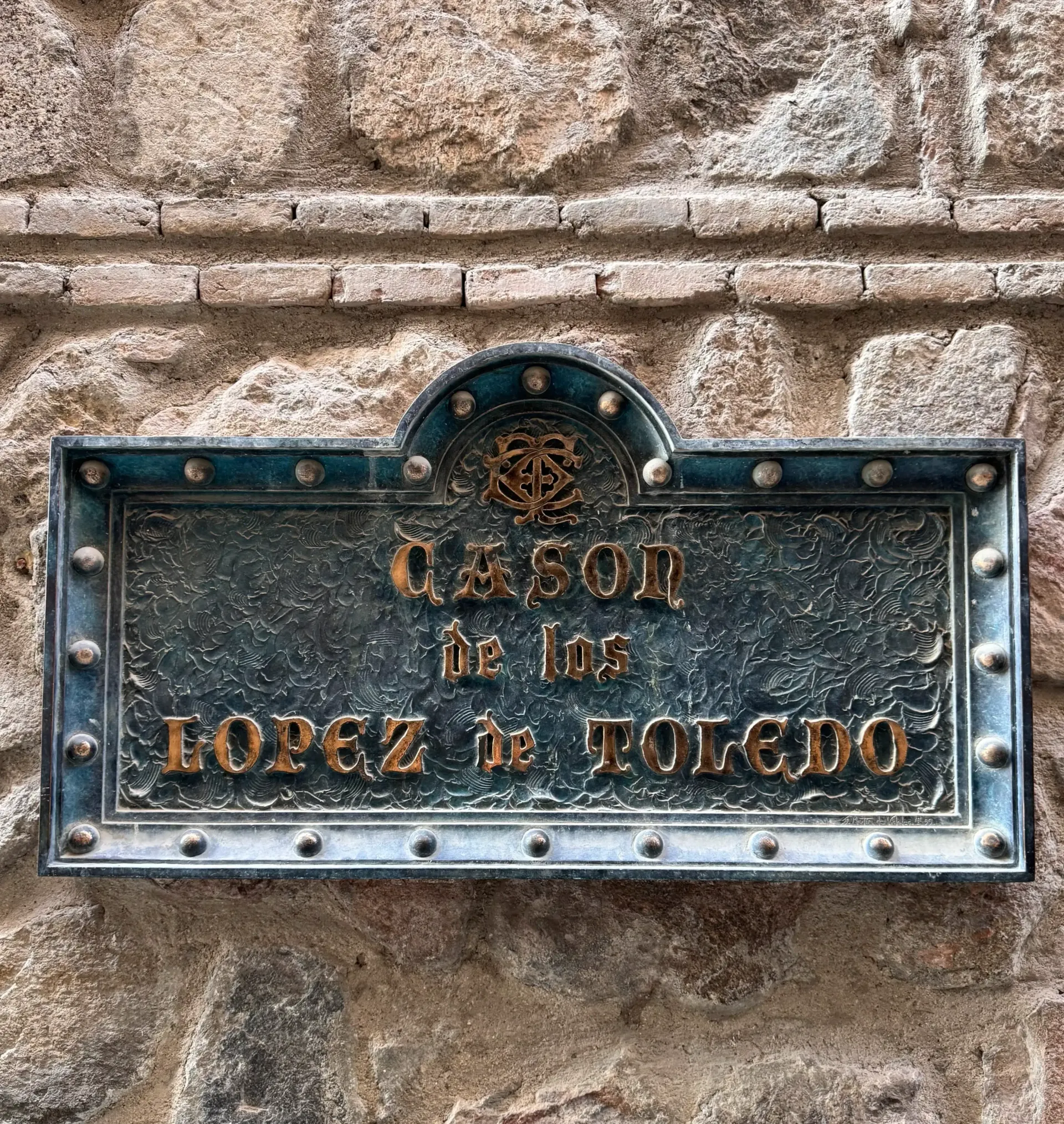 Una placa rectangular de color verde oscuro, remachada en un muro de piedra, reza 