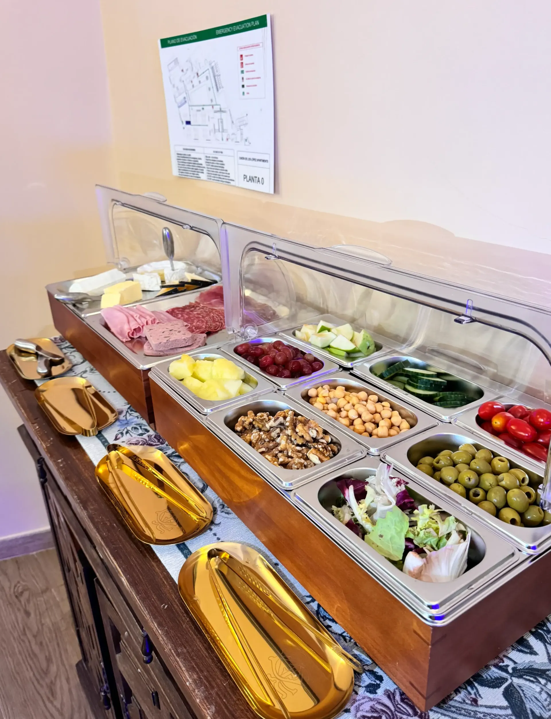 Una barra de ensaladas tipo bufé con diversos ingredientes como aceitunas, garbanzos y quesos, dispuestos en recipientes metálicos sobre un mostrador.