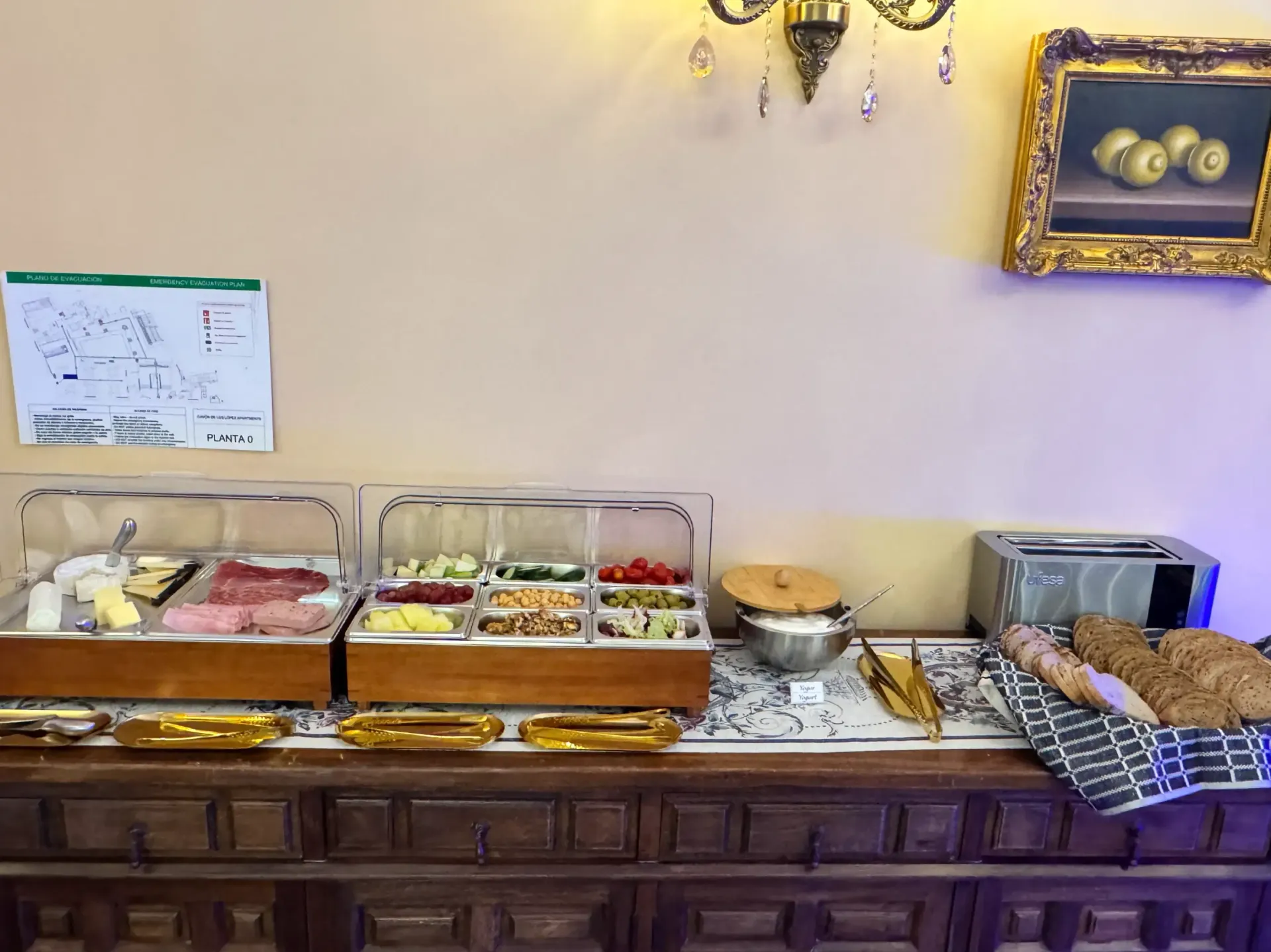Un bufé de desayuno de hotel con varios embutidos, quesos, verduras y pan dispuestos en una mesa auxiliar de madera.