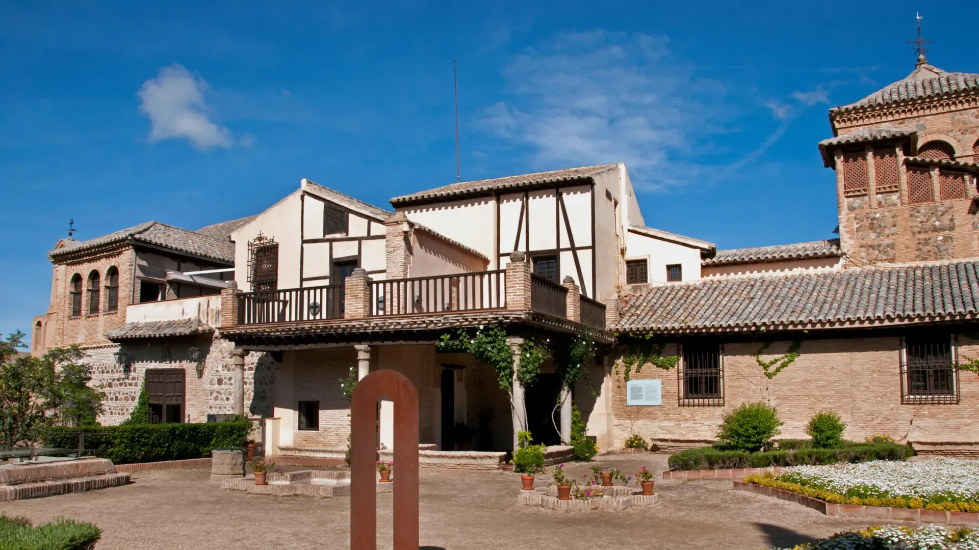 Un edificio histórico de estilo rústico con muros de piedra, vigas de madera, tejados de tejas, un balcón y un jardín bajo un cielo azul brillante.