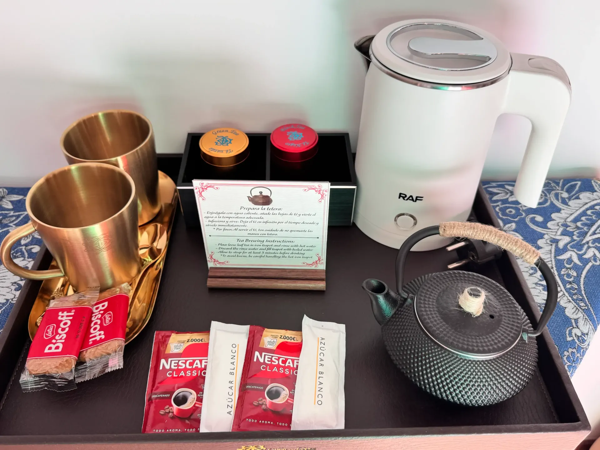 Una bandeja de hotel con dos tazas de latón, un hervidor eléctrico blanco, una tetera pequeña oscura, sobres de café instantáneo y aperitivos.