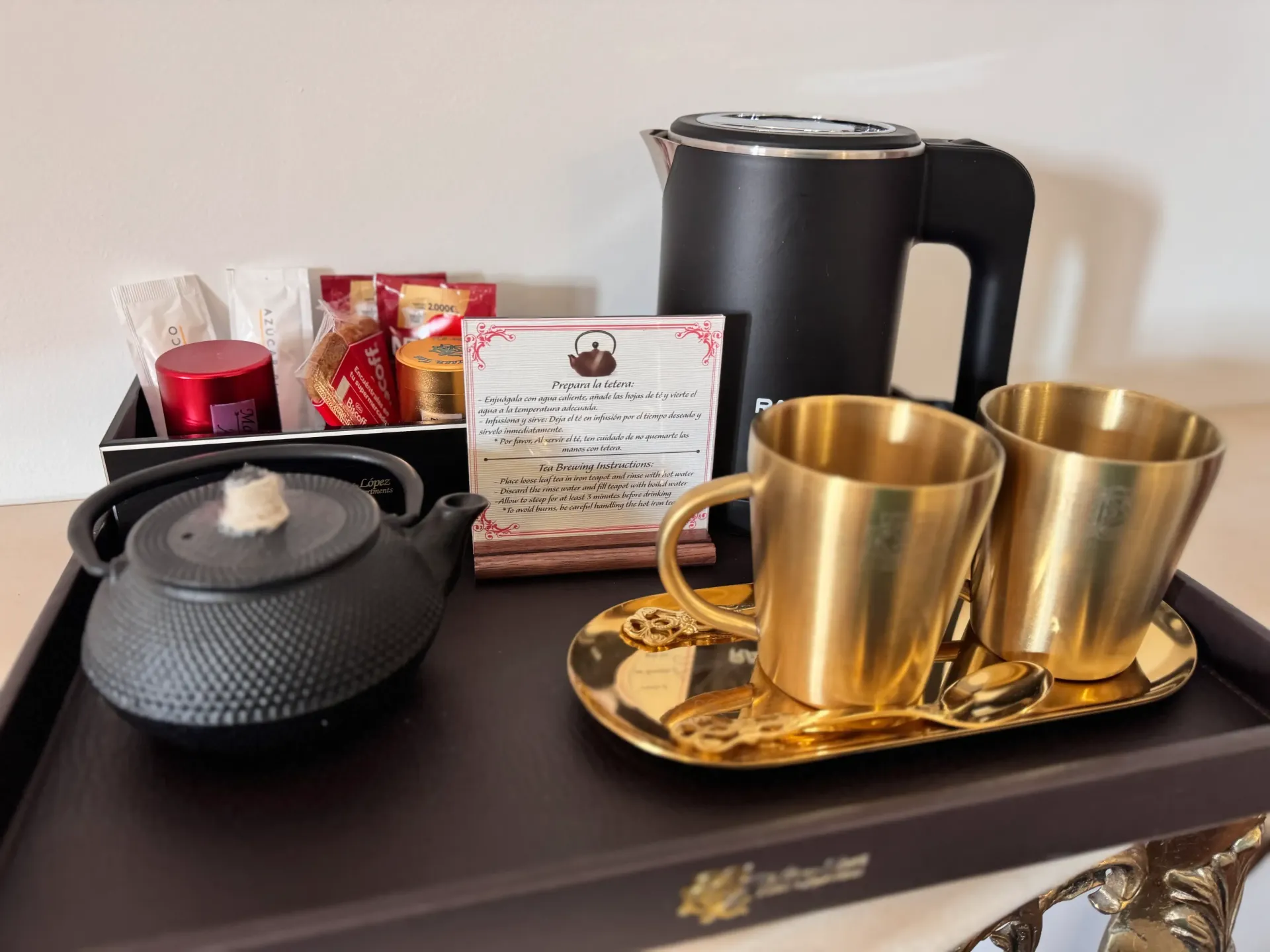 Un juego de té y café en una bandeja, que incluye un hervidor eléctrico negro, una tetera de hierro fundido y dos tazas de color dorado.