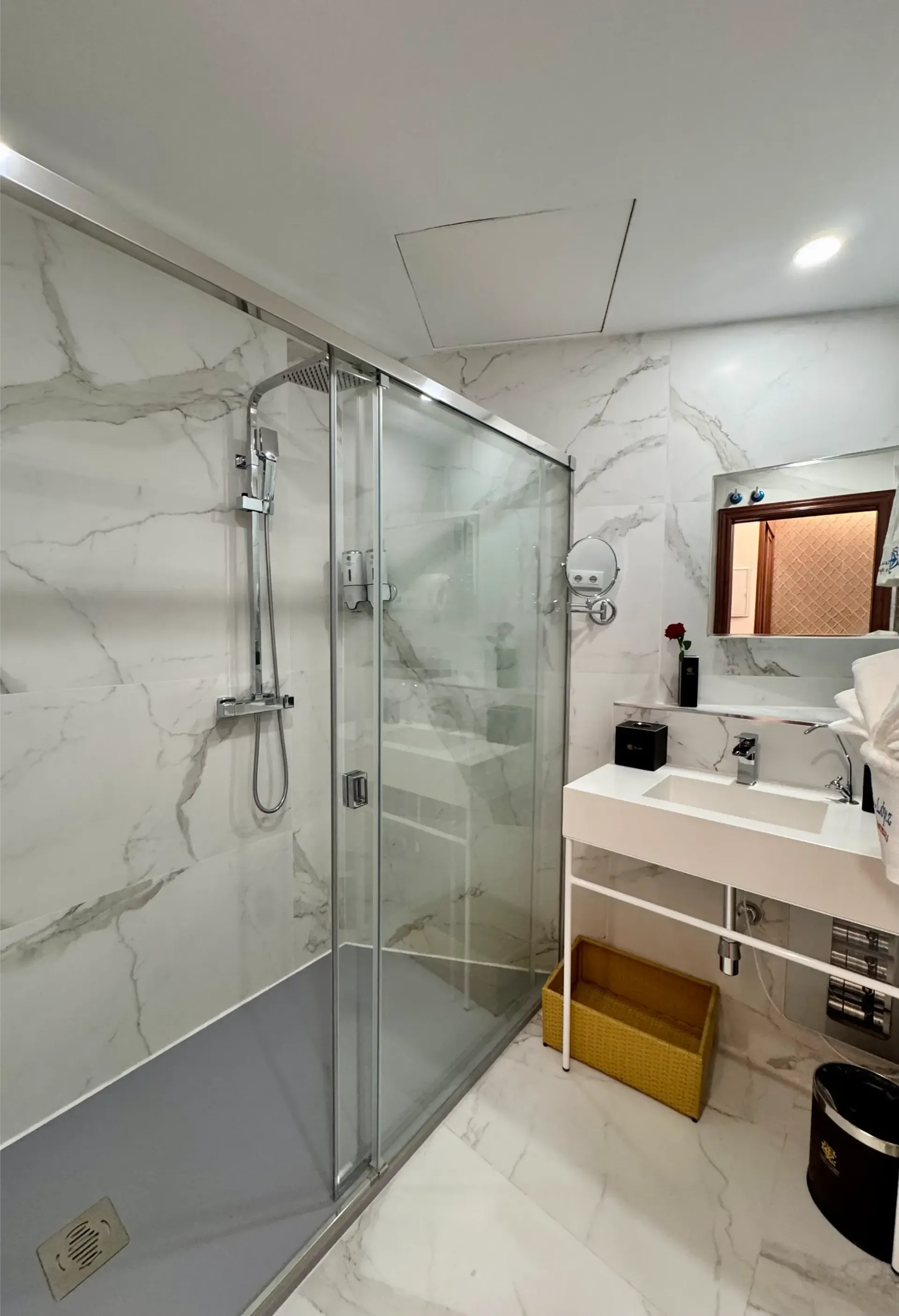 Baño moderno con ducha acristalada, paredes con diseño de mármol, lavabo blanco y cesta de almacenamiento amarilla.